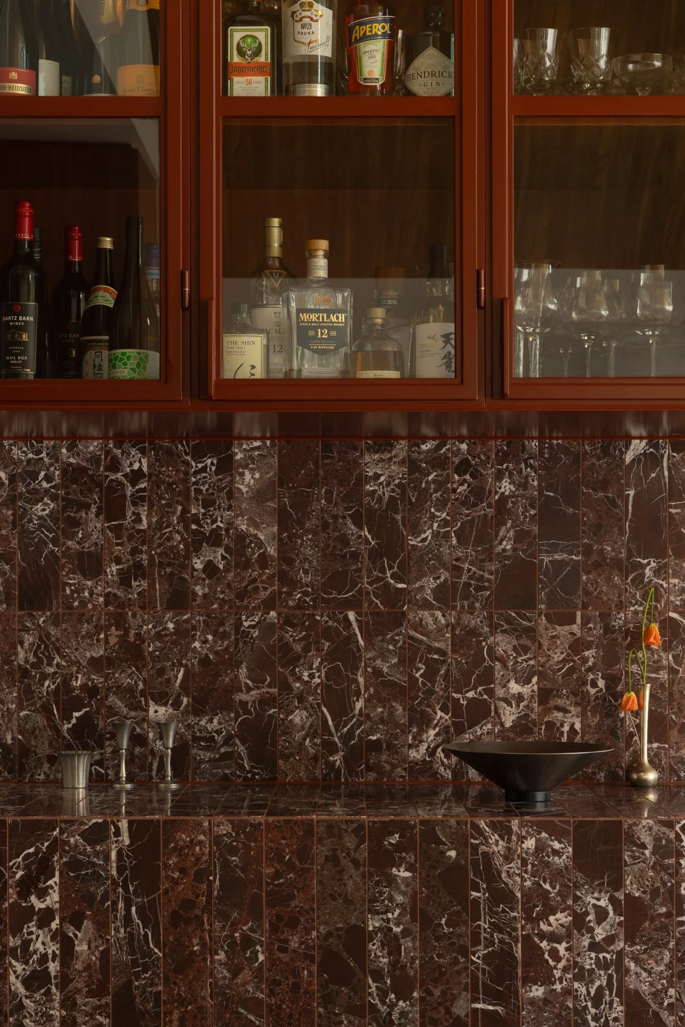 A bar cabinet with bottles of alcohol, glasses, and bar accessories behind glass doors. The cabinet is mounted on a dark, marble-tiled wall with a matching marble counter below.
