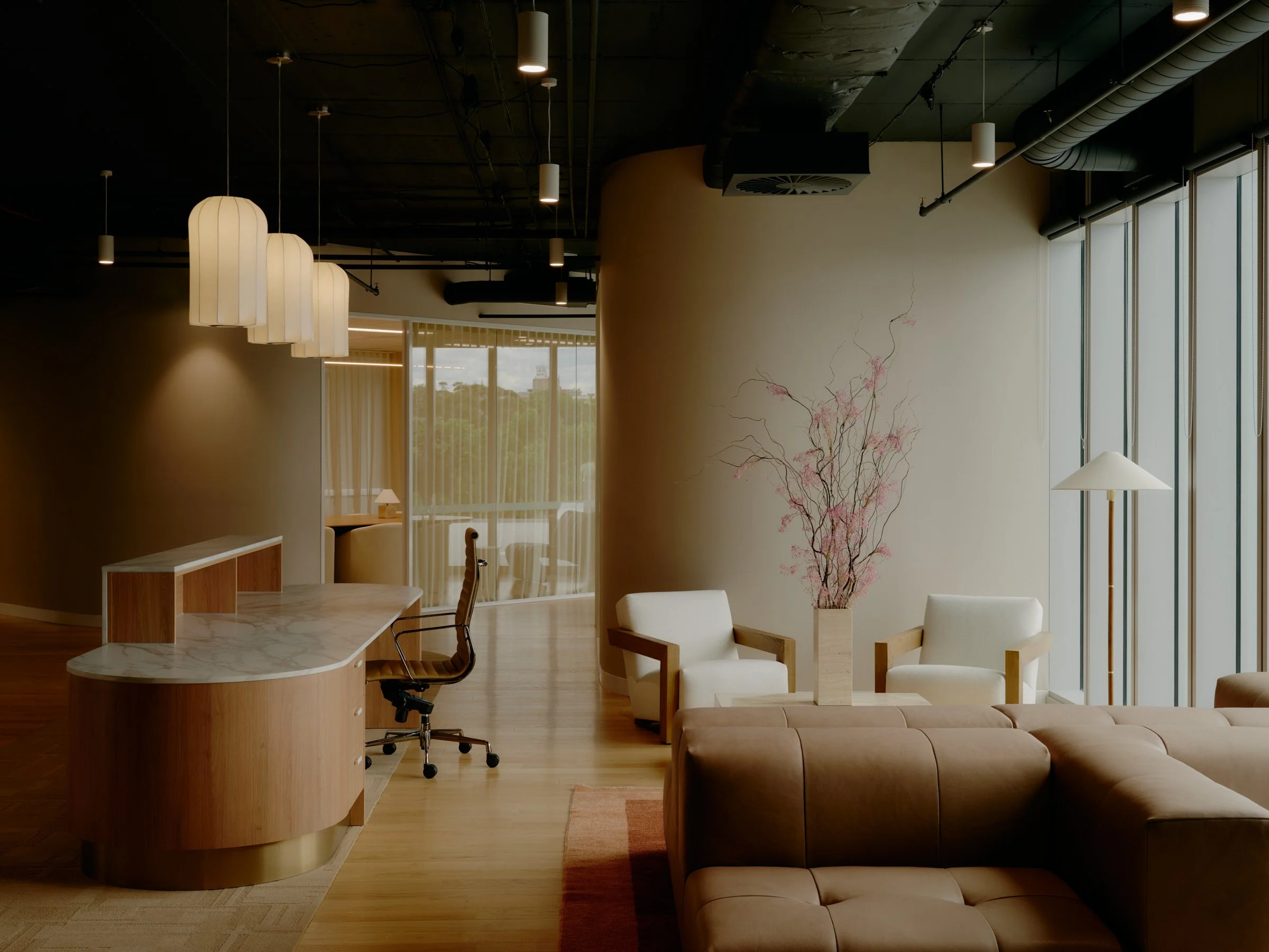 Modern interior lounge area with large windows, white armchairs, a brown leather sofa, a floor lamp, a large vase with pink flowering branches, and a wooden desk with an office chair.