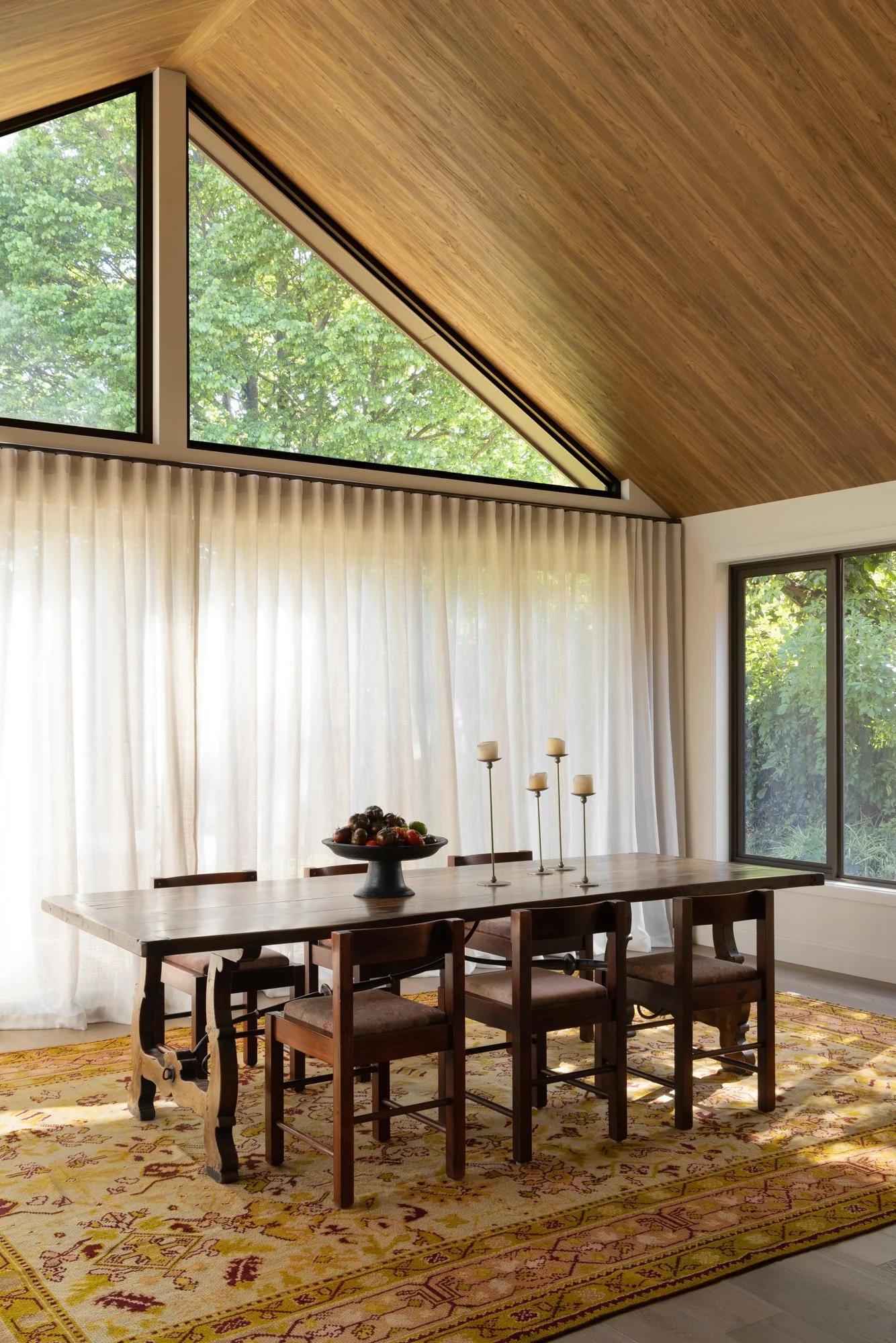 Interior of a room with a wooden sloped ceiling, large windows showing green trees outside, cream curtains, a wooden dining table, chairs, a bowl of fruit, candle holders, and a patterned area rug.