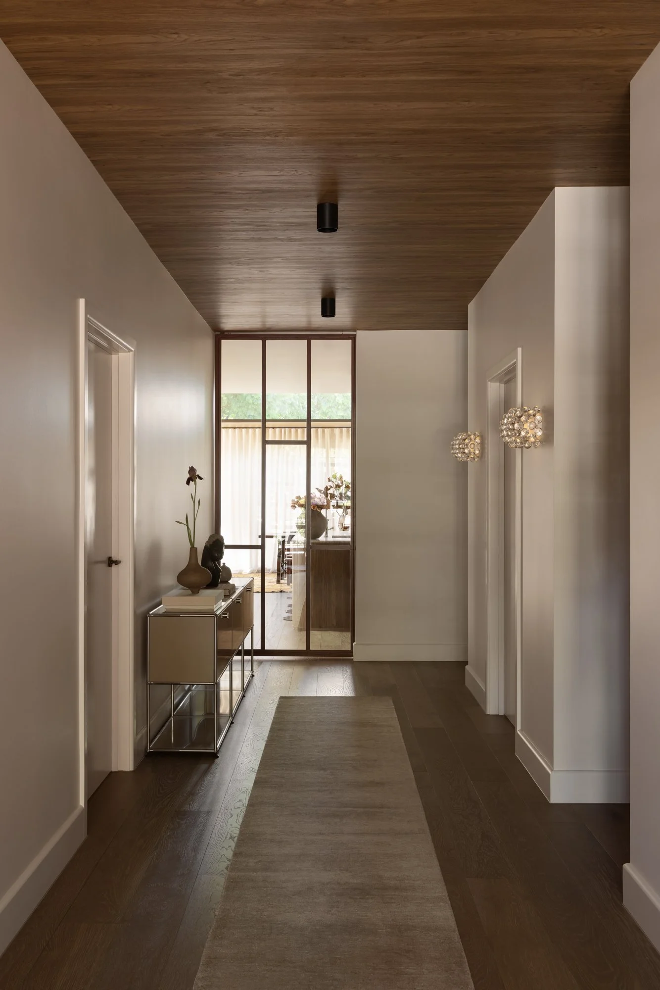 A modern hallway with a wooden ceiling, white walls, and dark wooden floors. There is a small console table with decorative items and a tall vase with a flower. Beyond the hallway, there is a room with large windows, sheer curtains, and a potted plan