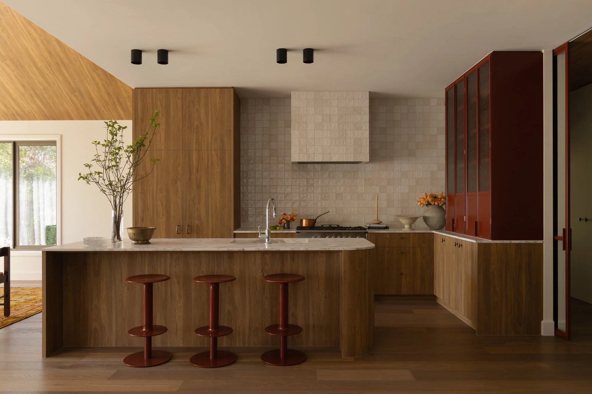 Modern kitchen with wooden cabinetry, a marble island with two red bar stools, beige tiled backsplash, and decorative plants and bowls