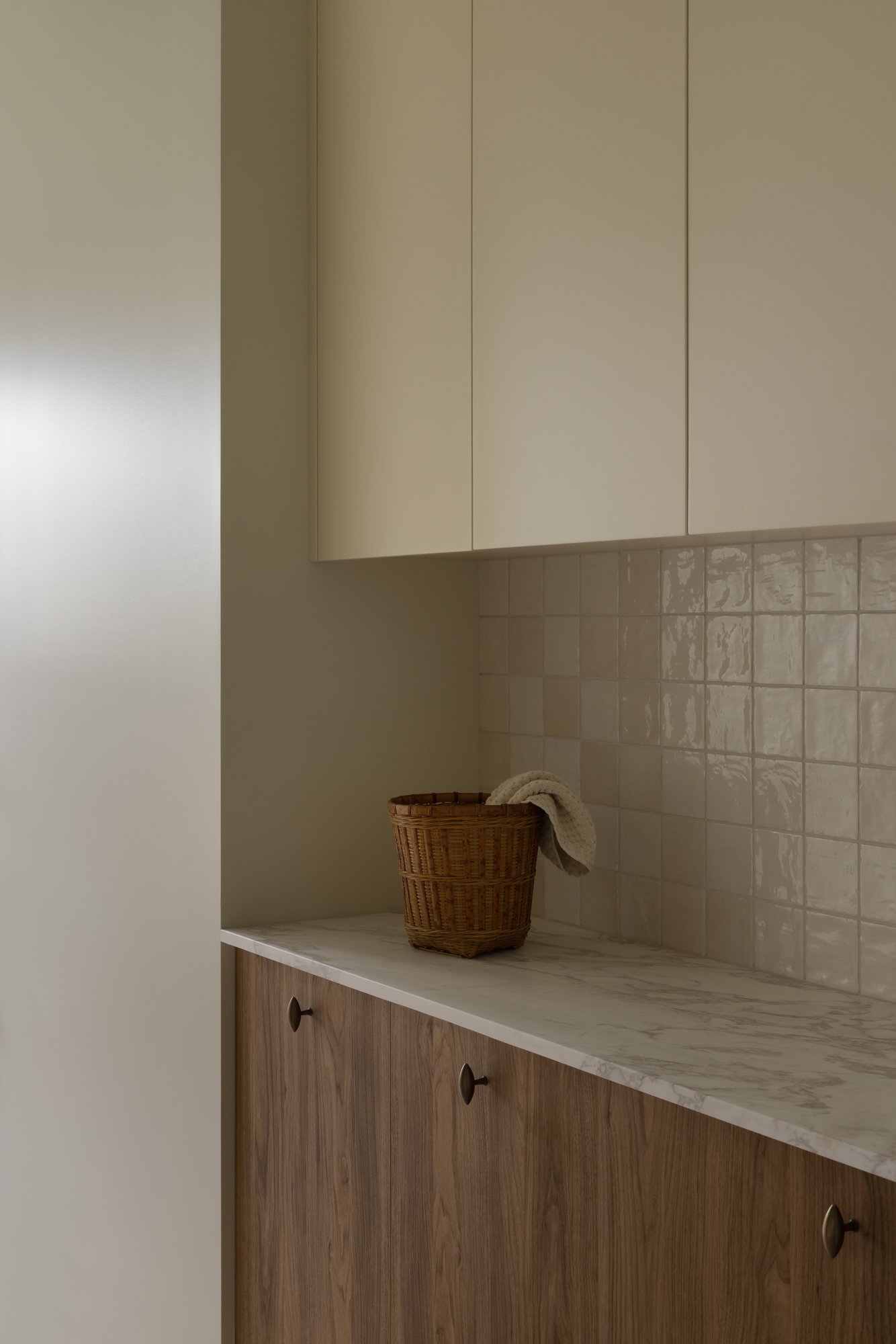 Bathroom countertop with a basket and towel, beige cabinets, tiled wall.