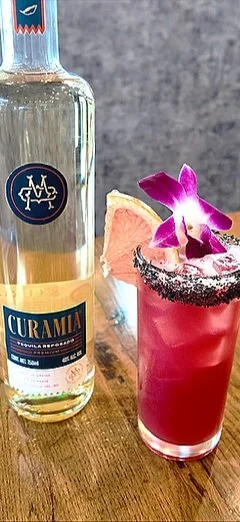 A turquoise bottled spirit labeled 'Curaçao' next to a pink cocktail garnished with a purple orchid, a pineapple slice, and black salt rim on a wooden surface.