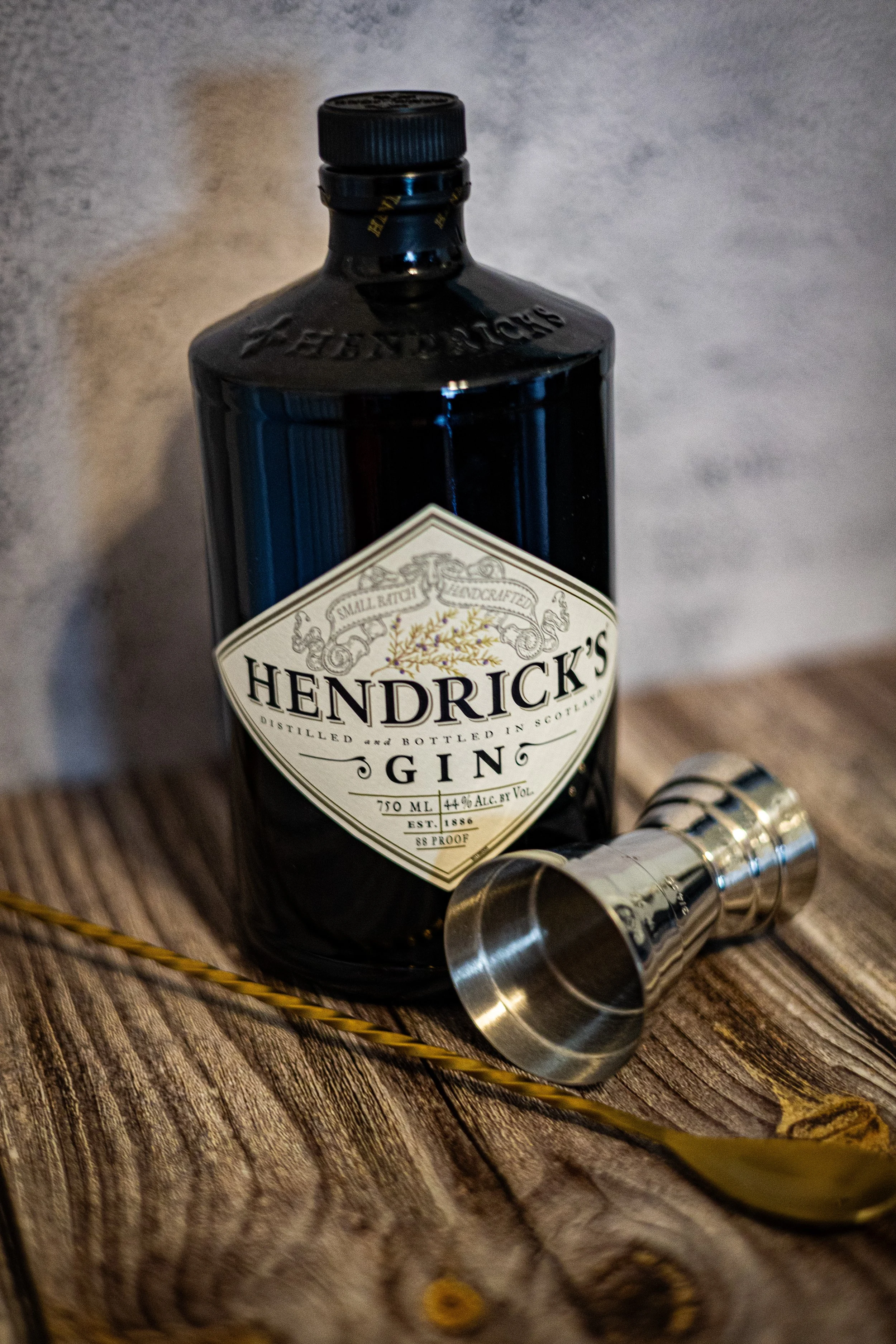A bottle of Hendrick's gin, a metal jigger, and a long yellow spoon on a rustic wooden surface.