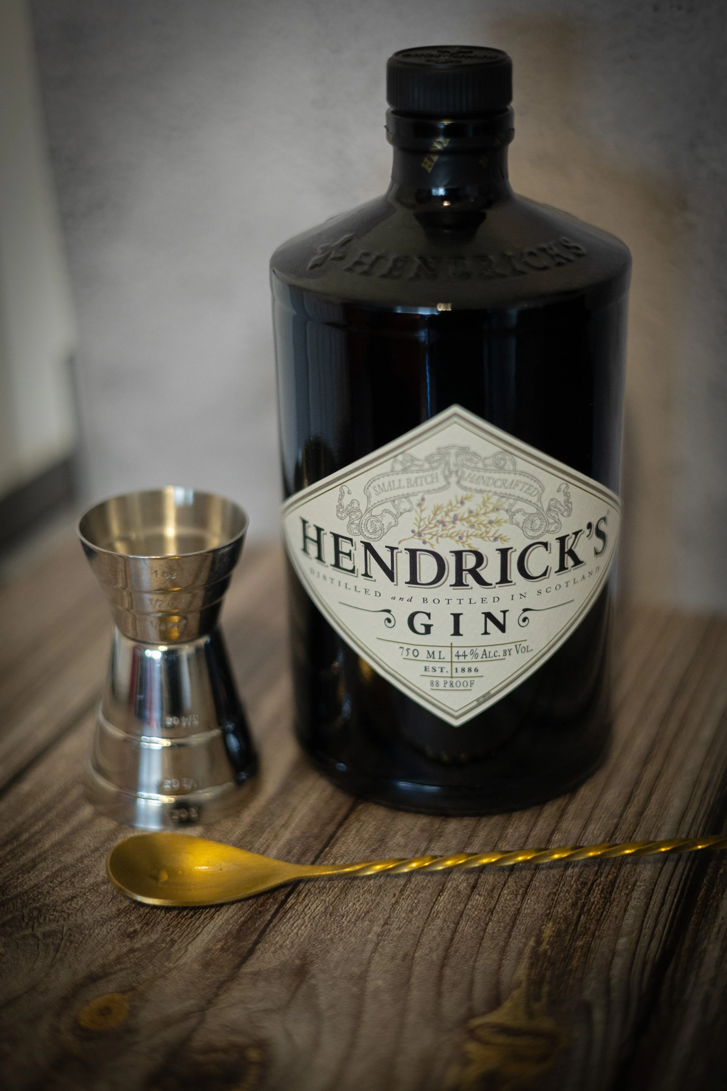 A large bottle of Hendrick's gin, a small metallic jigger, and a gold spoon on a wooden surface.