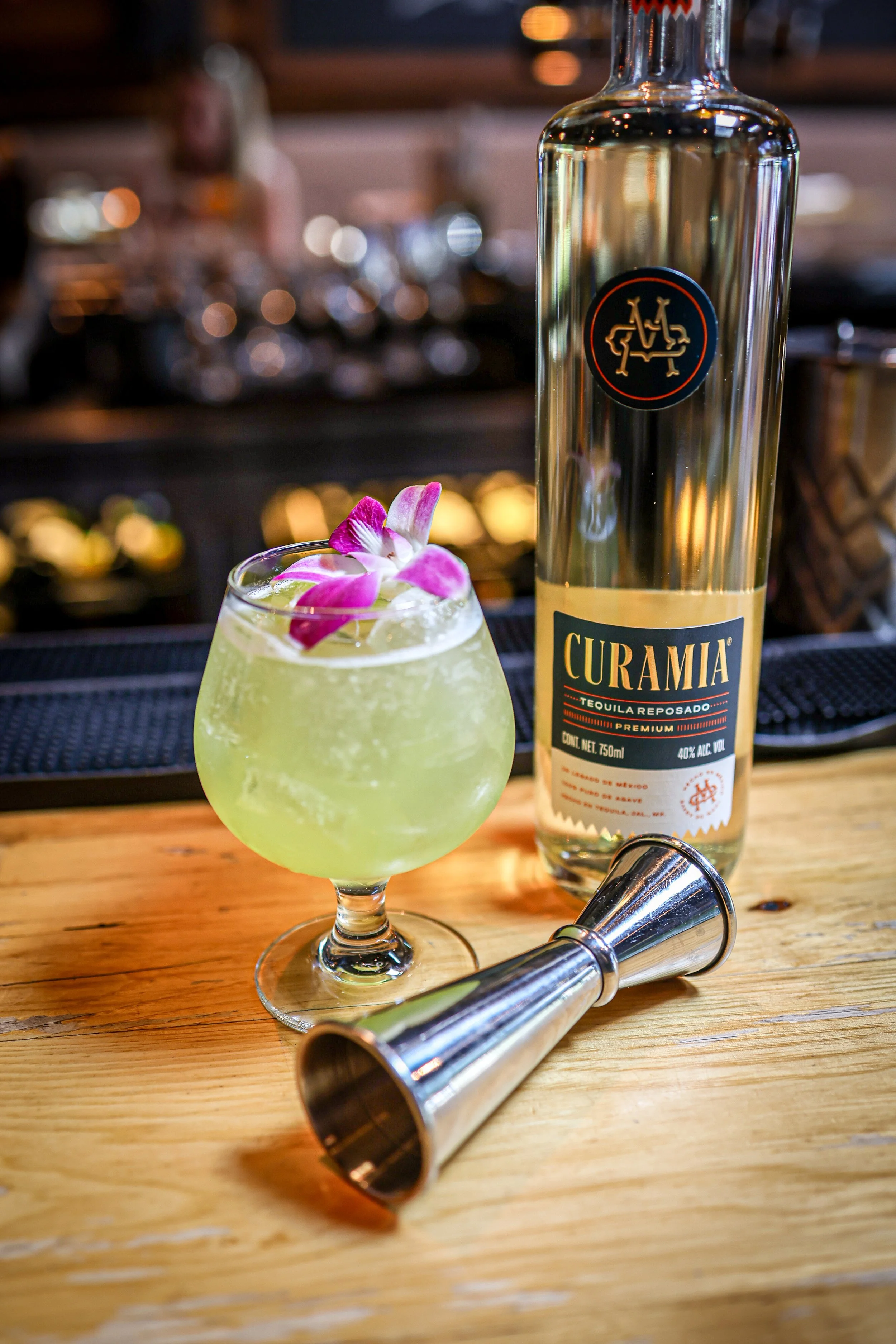 A cocktail in a rounded glass garnished with pink and white flowers, a bottle of Curamia tequila, a stainless steel jigger, on a wooden bar counter.