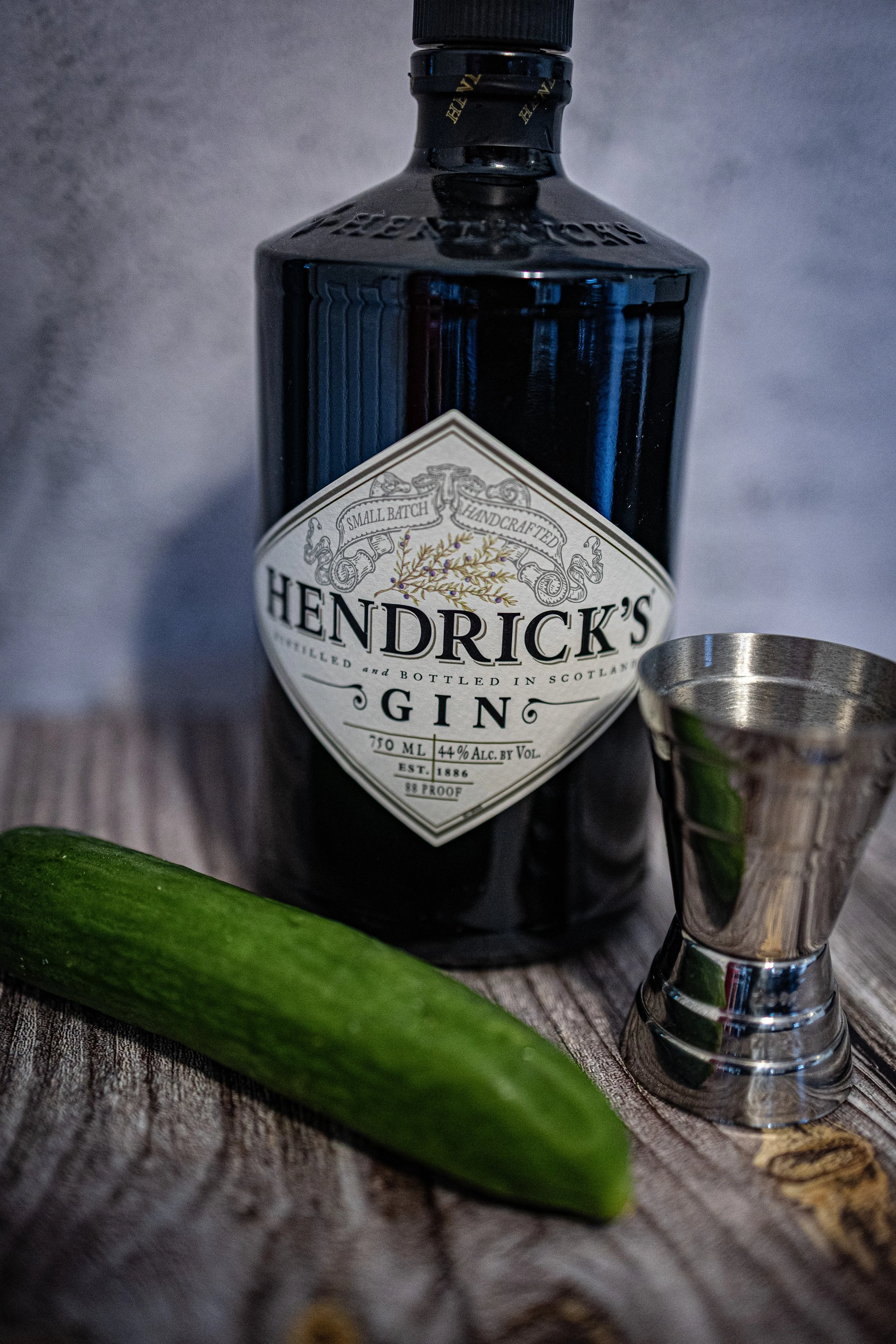 A bottle of Hendrick's Gin with a silver cocktail jigger and a cucumber on a rustic wooden surface.
