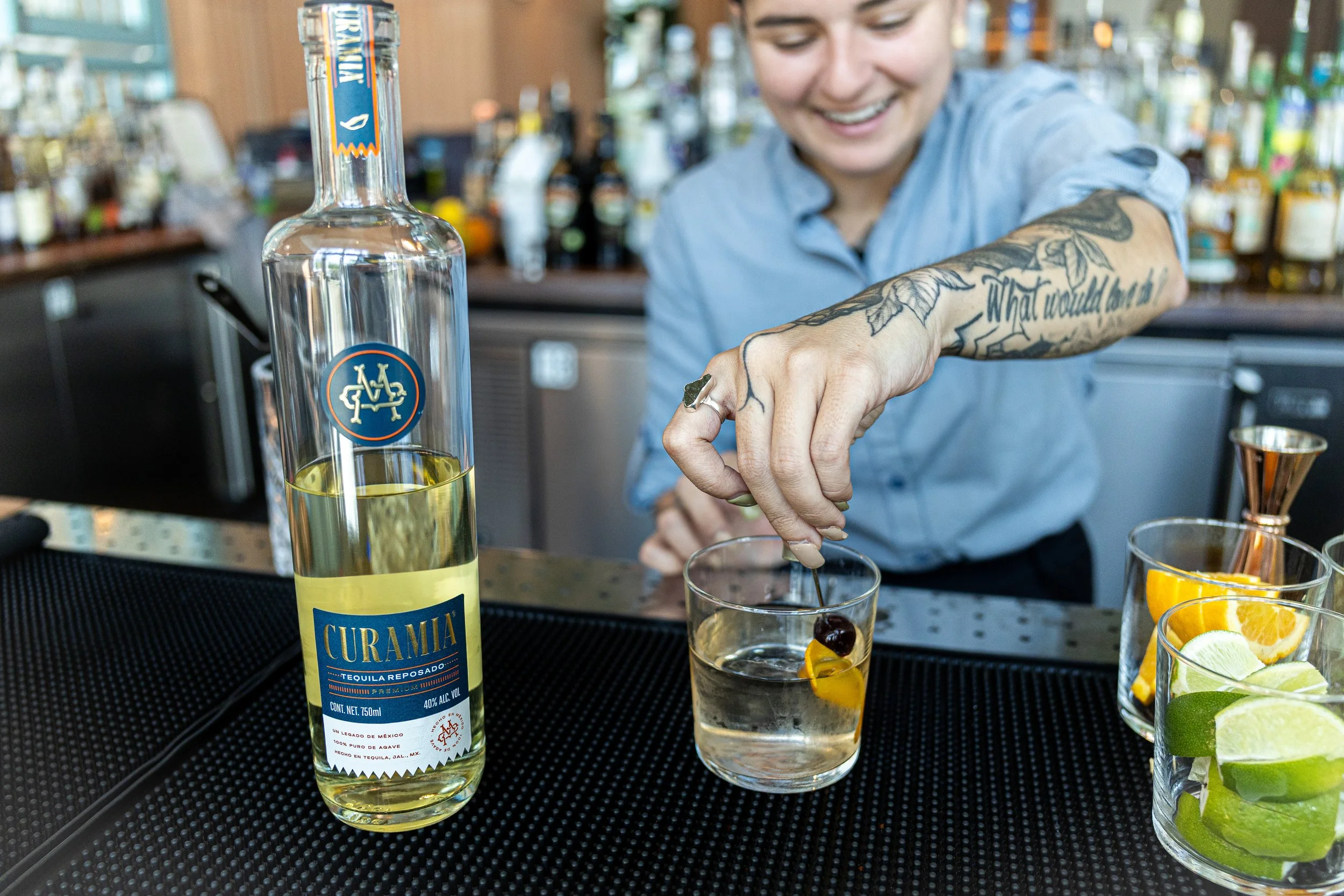 Bartender placing an olive into a glass of clear liquid with lemon peel, with a bottle of tequila and citrus slices on the bar.
