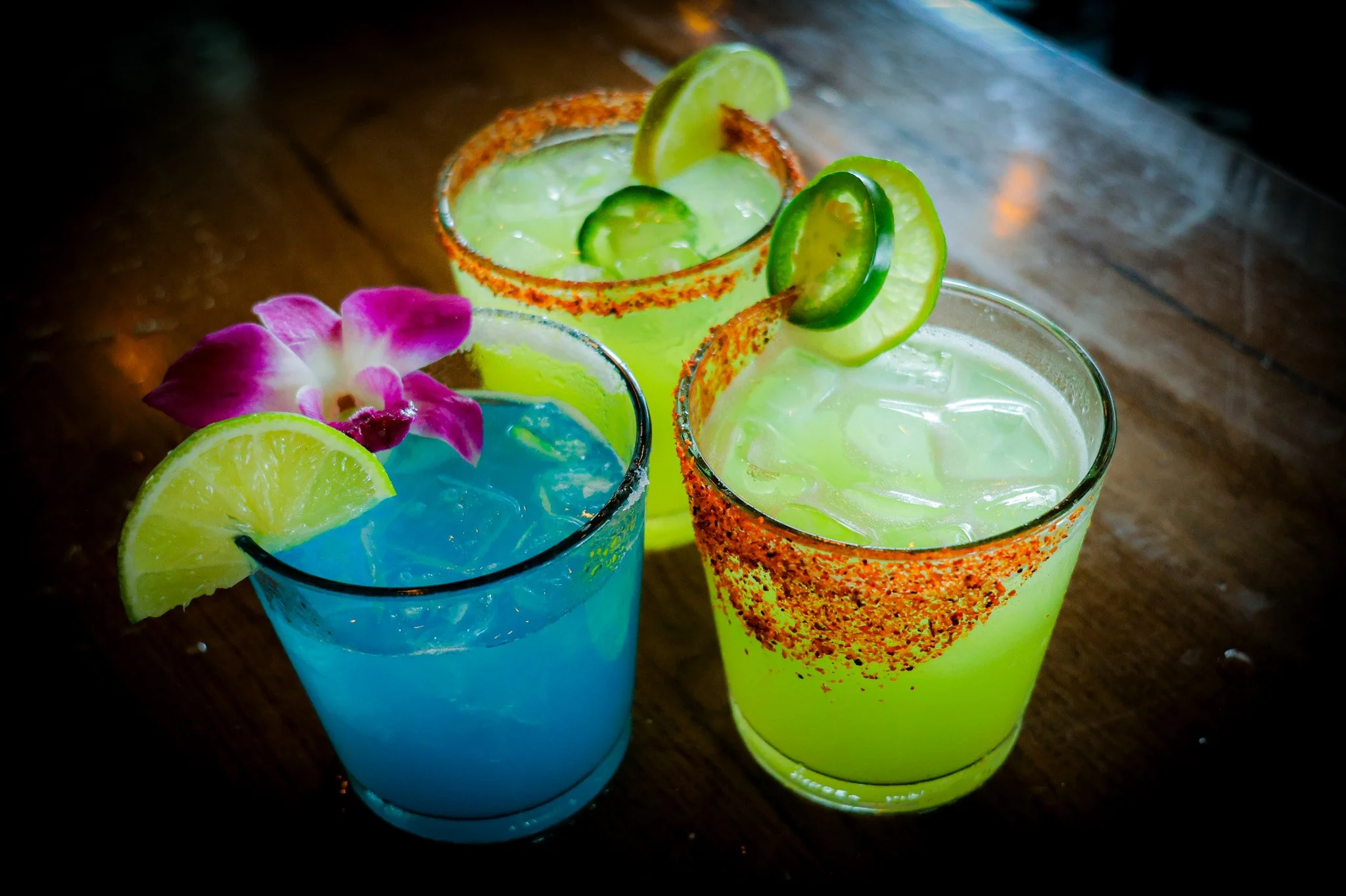 Three colorful cocktails on a wooden table, garnished with lime slices, cucumbers, and a pink flower.