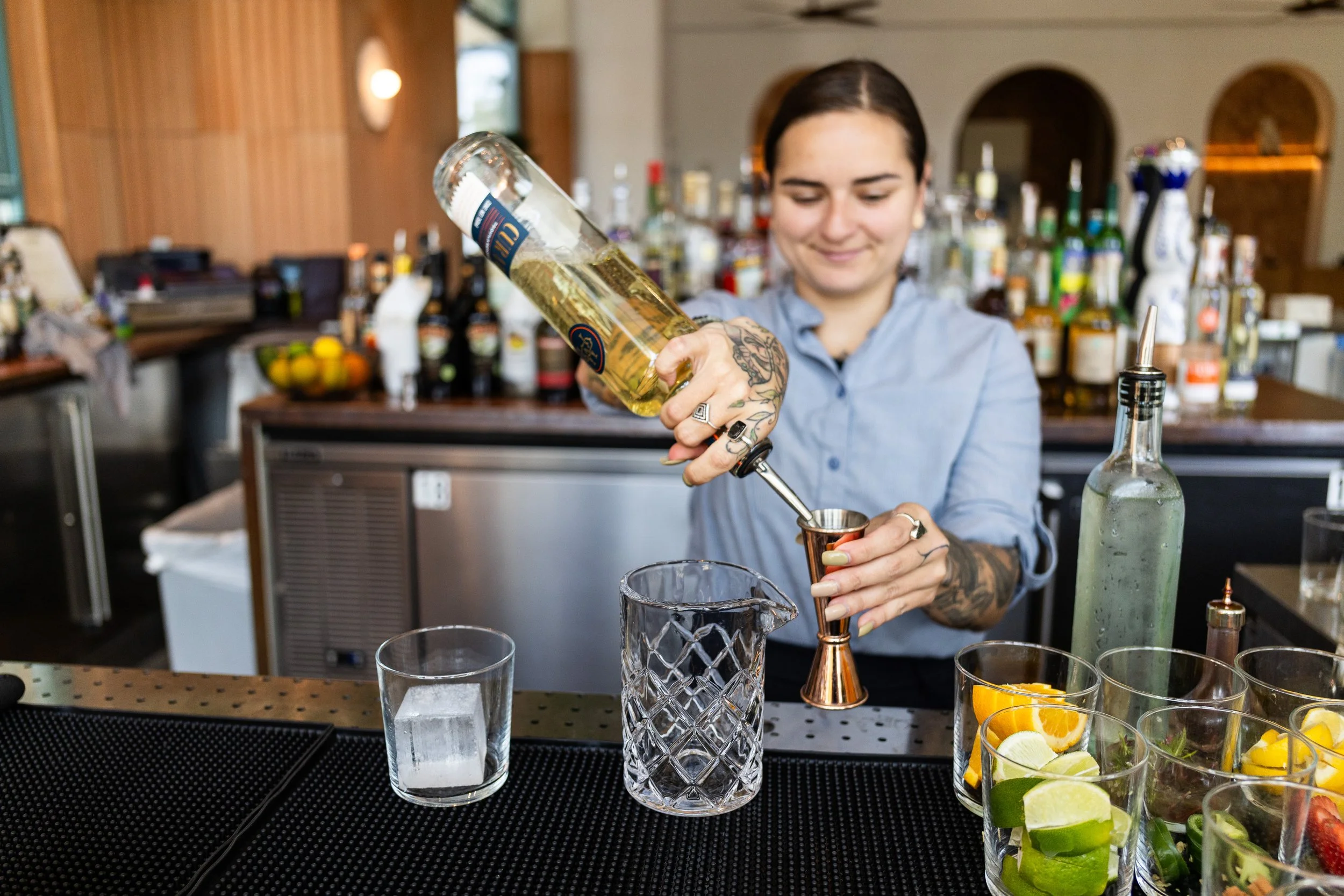 A female bartender with tattoos preparing a cocktail at a bar, pouring liquor into a jigger over a glass pitcher, with various fruits and liquor bottles in the background.