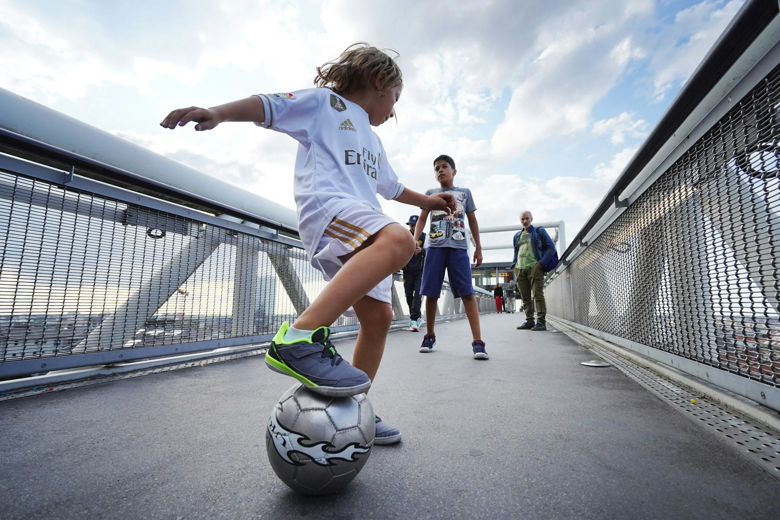 A young girl in a white soccer uniform balancing on a soccer ball on a bridge, with two boys and an adult in the background.