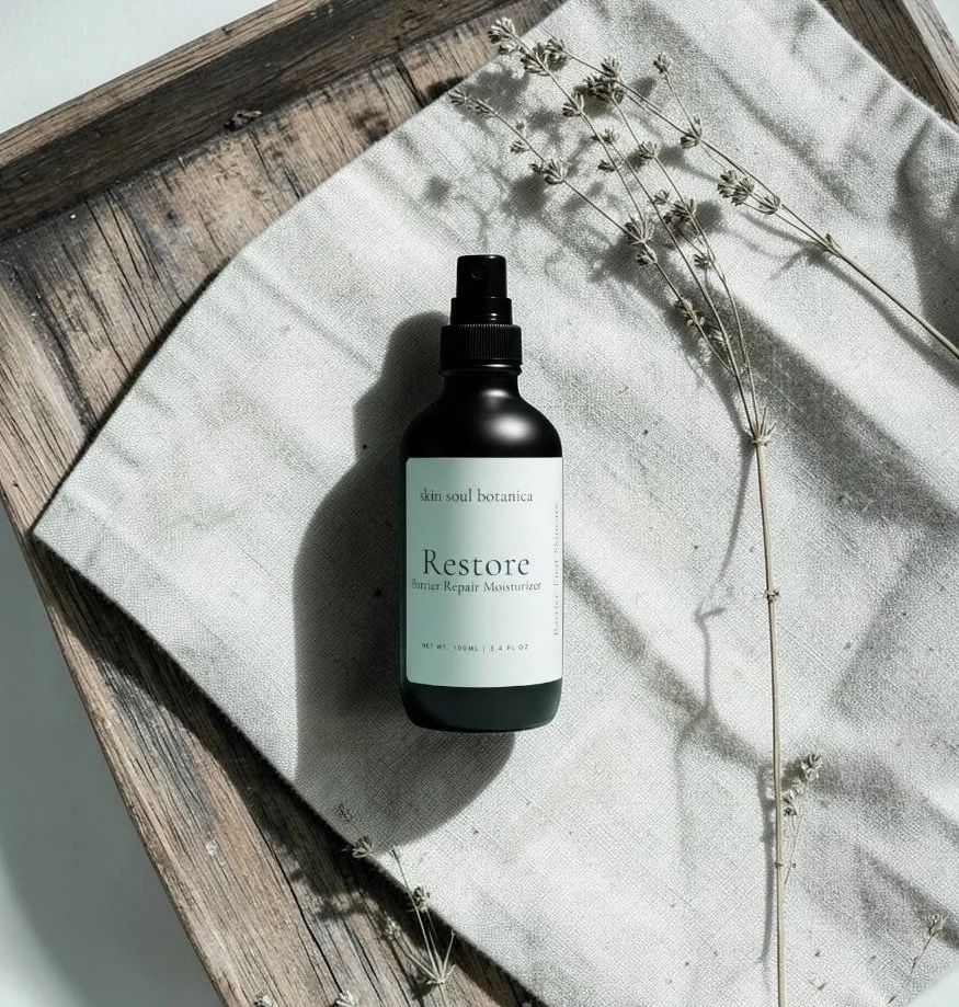 A black bottle of Skin Soul Botanica Restore Barrier Repair Moisturizer spray on a white cloth with dried flowers, on a rustic wooden surface.