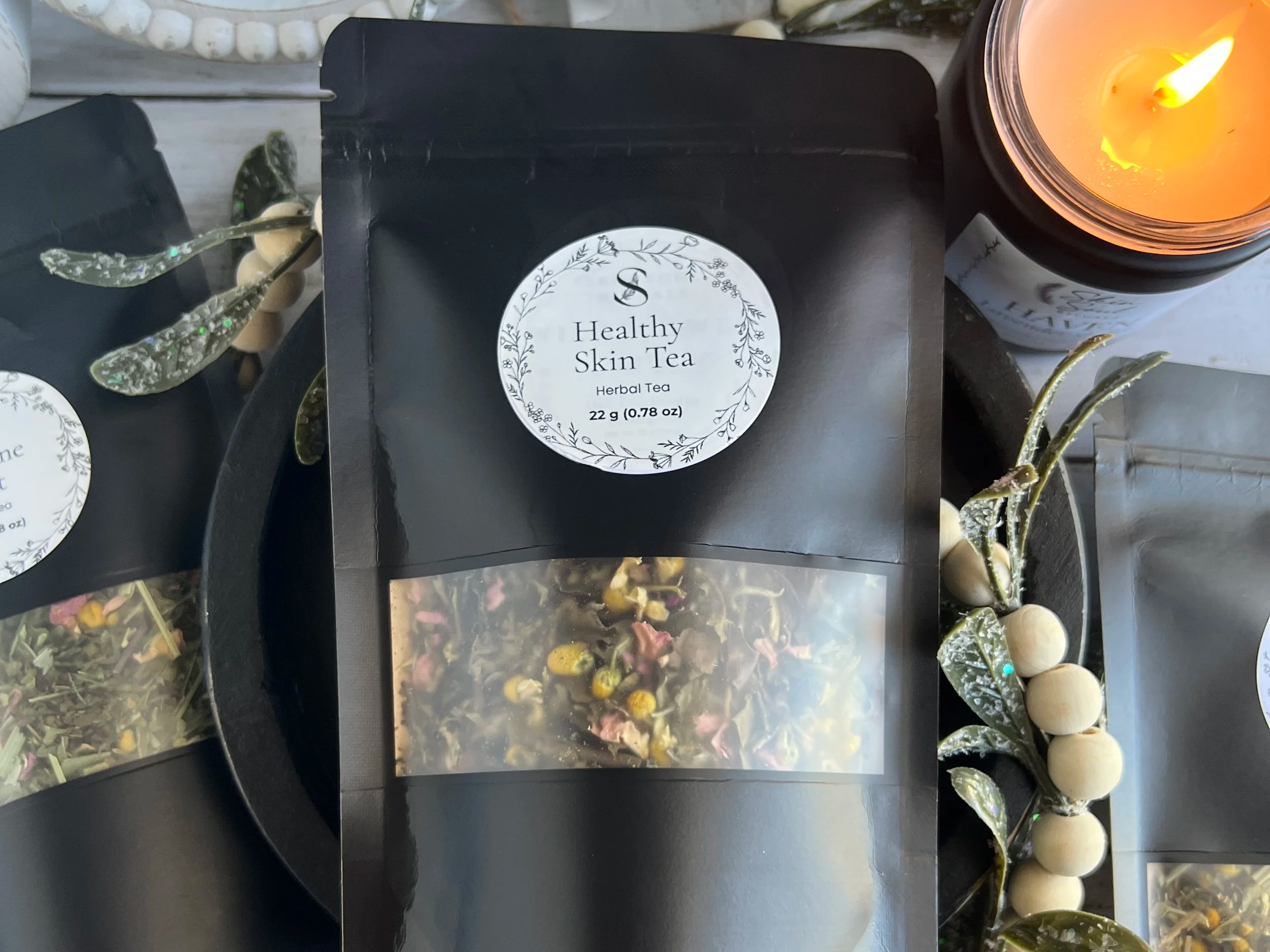 Close-up of herbal teas, candles, and decorative items on a table. The herbal tea package labeled 'Healthy Skin Tea' is in the center with a black and white floral design. A lit candle is on the right side, emitting a warm orange glow. There are also other herbal tea bags and decorative metallic leaves with glitter around the package.