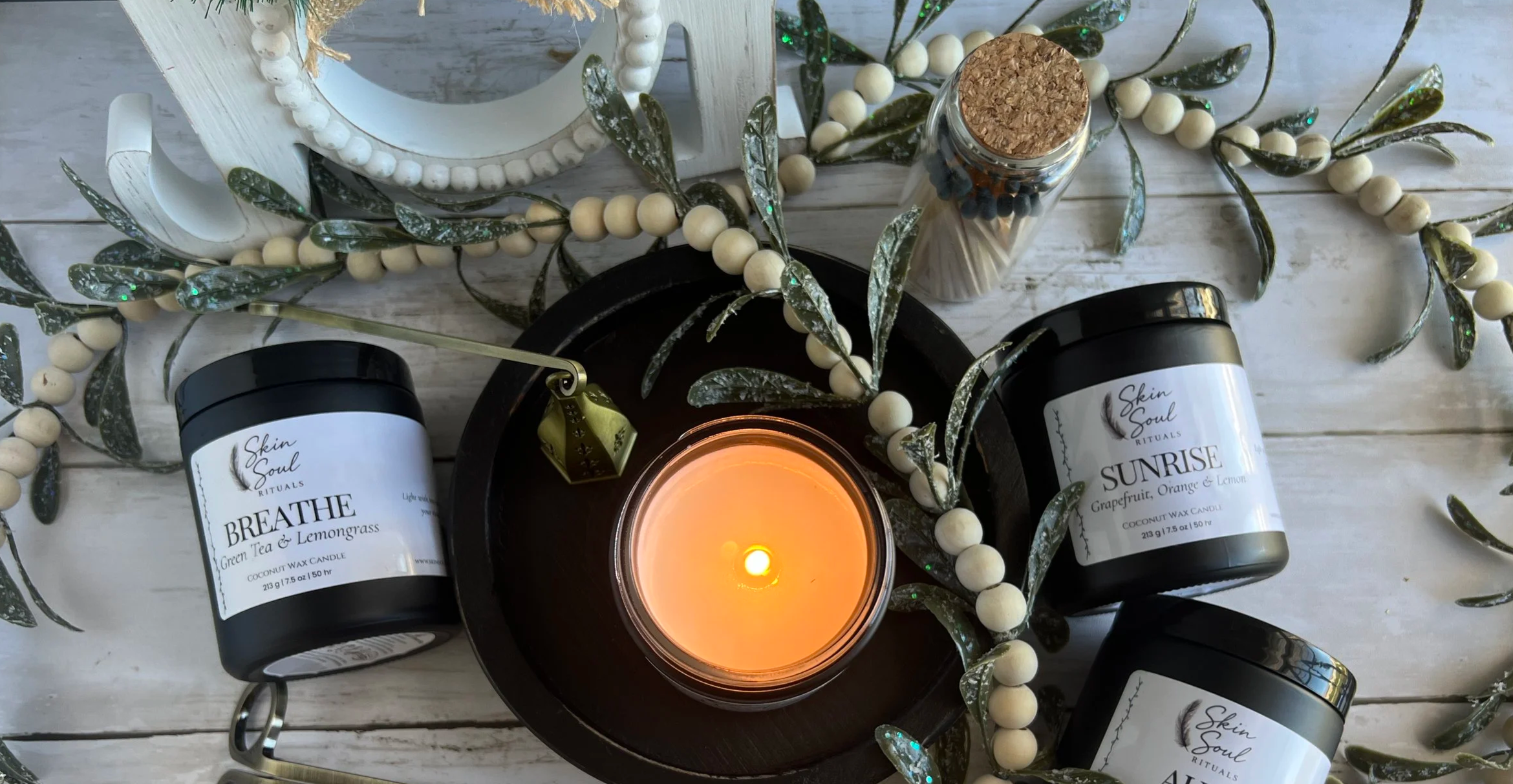 A lit candle surrounded by decorative greenery, candles, and containers of Skin Soul Rituals candles labeled Breathe and Sunrise on a white wooden surface.