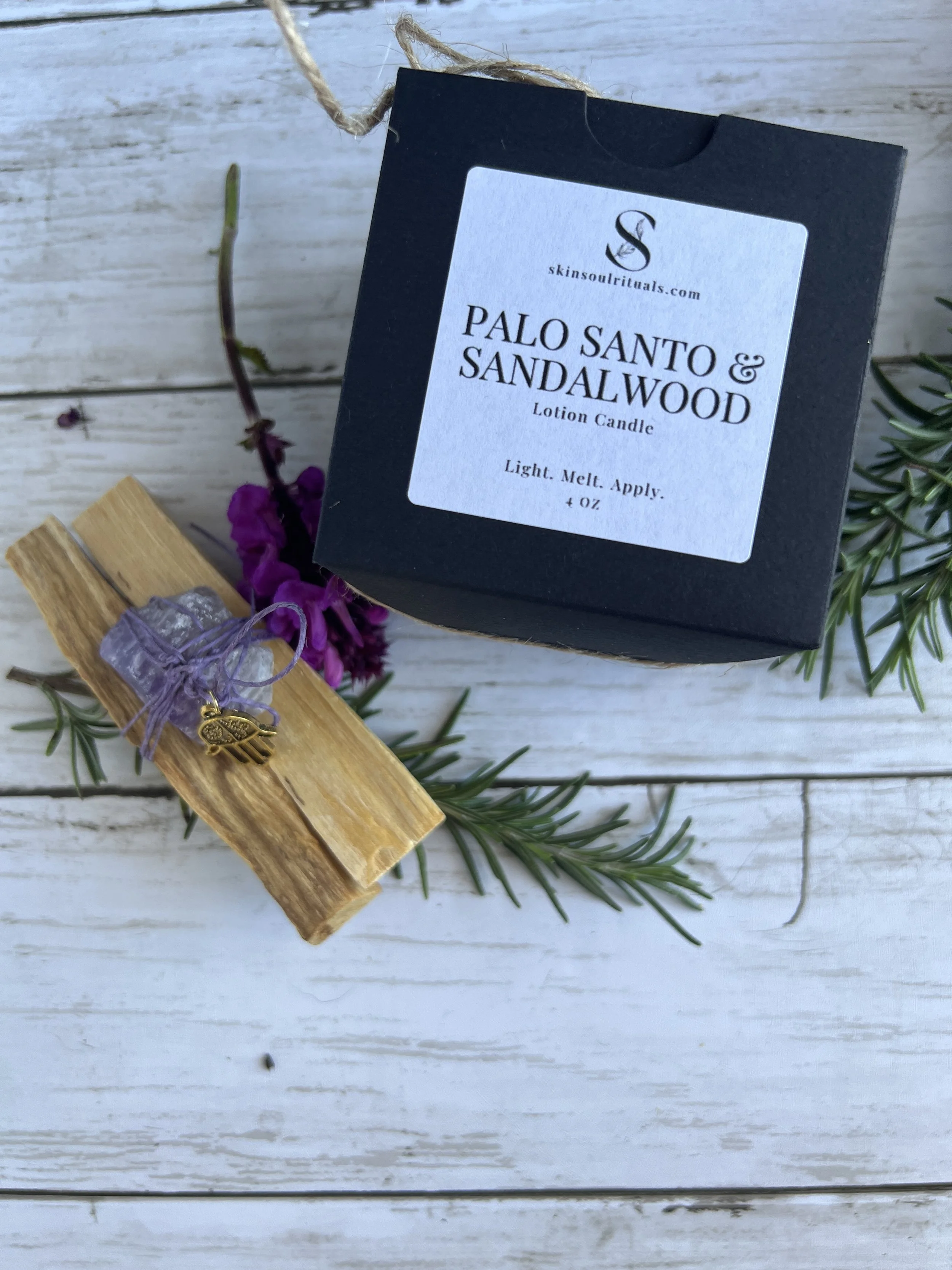 A black box labeled 'Palo Santo & Sandalwood Lotion Candle' with a description. . Both items are placed on a piece of greenery on a white wooden surface.