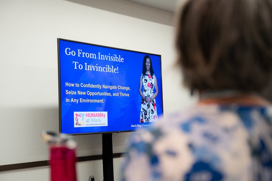 A woman watching a presentation on a large screen with blue background, titled 'Go From Invisible To Invincible!', with a woman in a floral dress smiling, and text about navigating change and seizing opportunities.