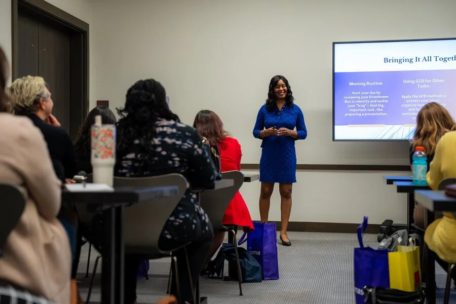 A woman in a blue dress giving a presentation to a seated audience in a conference room.