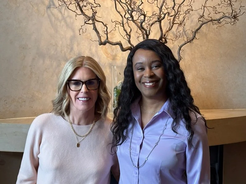 Two women smiling for a photo indoors, with a decorative tree sculpture and warm-colored wall behind them.
