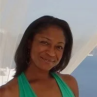 A woman with curly black hair smiling, wearing a green sleeveless top, standing in front of a light-colored curtain or drape.