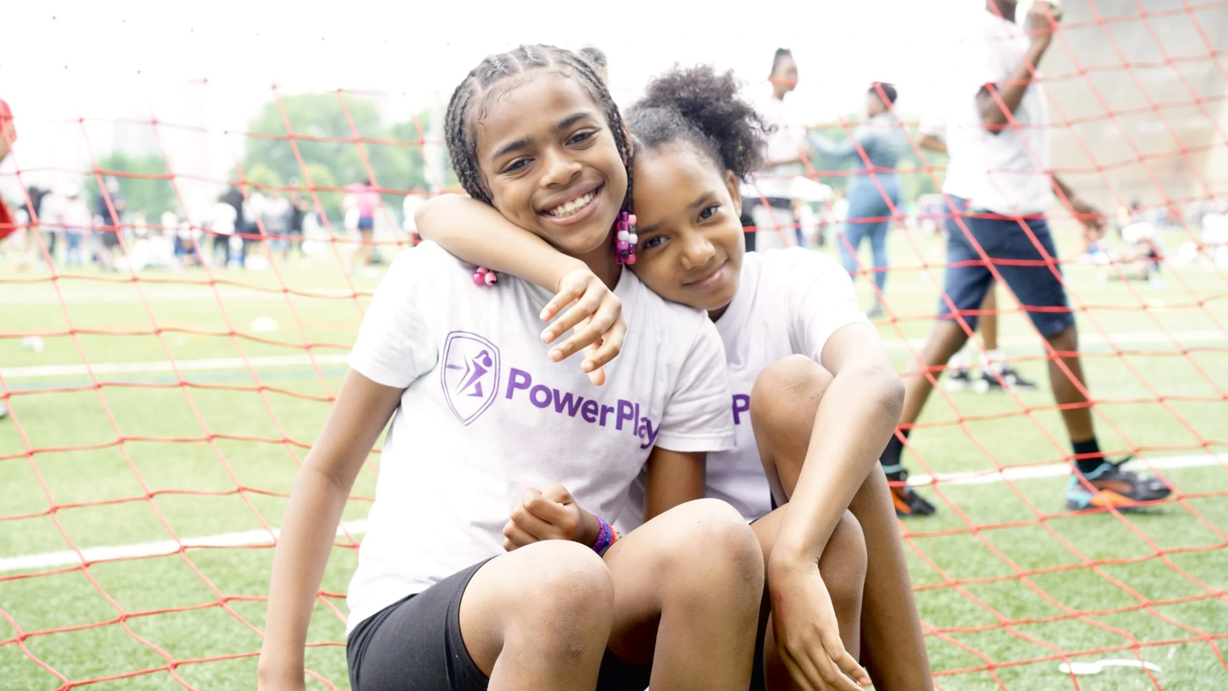 Two young girls sitting behind a red sports net at a field, smiling and hugging each other. They are wearing white t-shirts with a PowerPlay logo, and there are people in the background on a sports field.