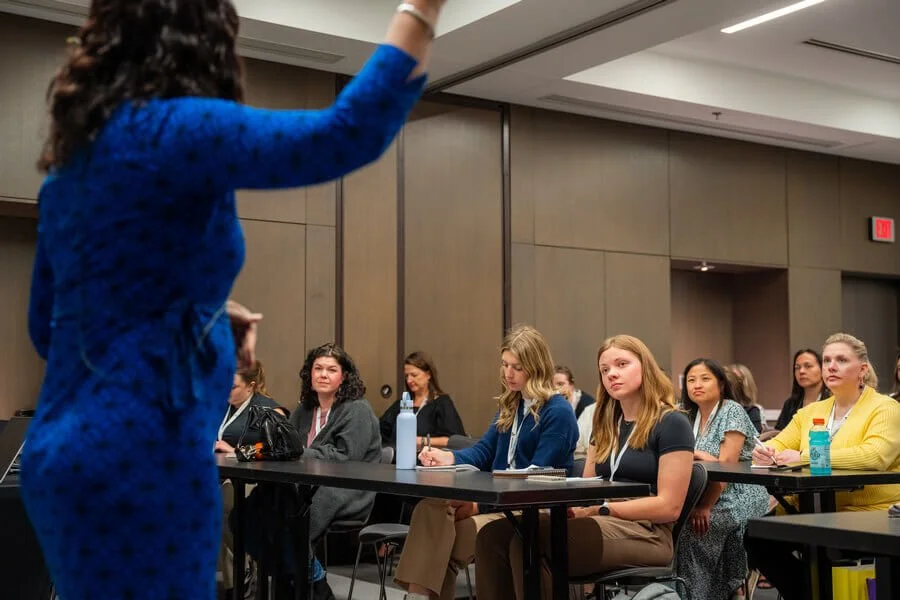 A woman in a blue dress is speaking or presenting to a group of women seated at tables in a conference room.