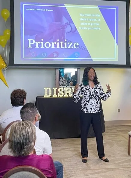 Woman giving a presentation on prioritizing goals to an audience at an indoor event, with a screen displaying the word 'Prioritize' and a quote about putting steps in place to achieve desired results, and large gold letters spelling 'DREAM' on a table behind her.