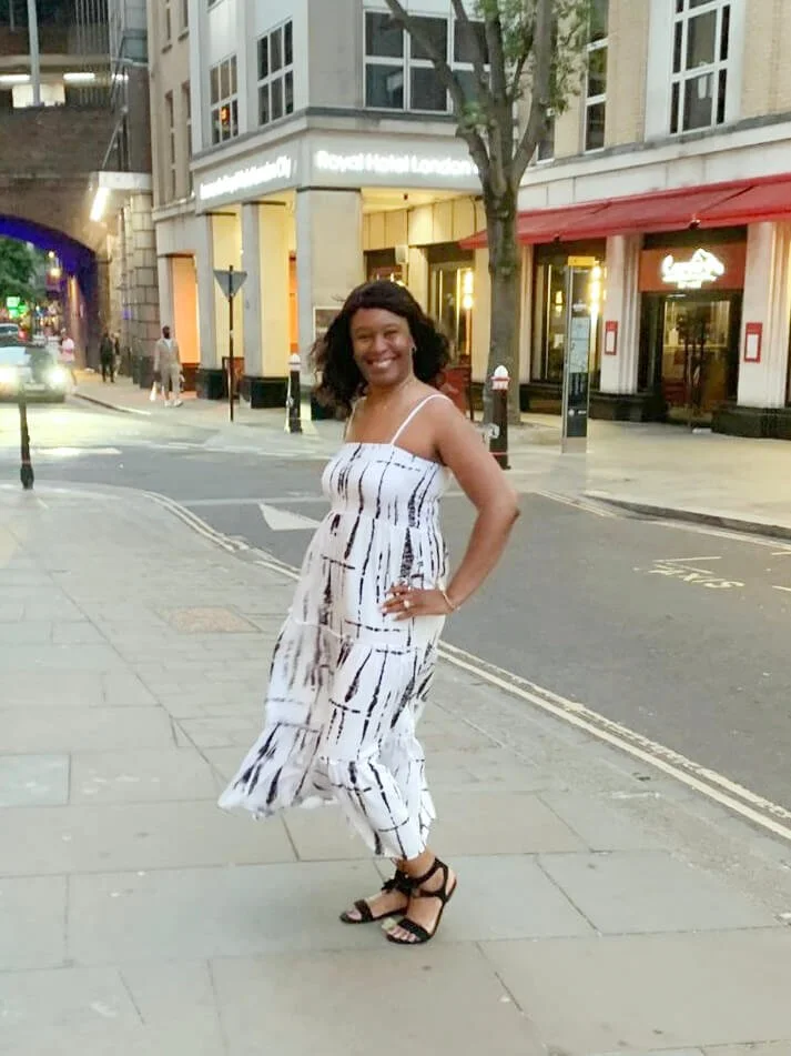 A woman in a white and black striped dress posing on a city sidewalk in front of a building with a sign reading 'Royal Hotel London'; she is smiling and wearing black sandals.