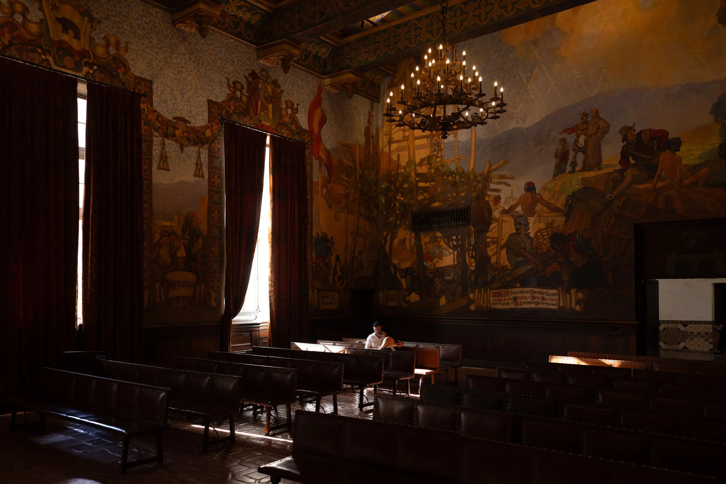 A man and woman sitting and courtroom in a dimly lit, historic room with dark wood furniture, large windows with heavy curtains, and a detailed mural painting on the wall depicting a historical scene with soldiers and a landscape.