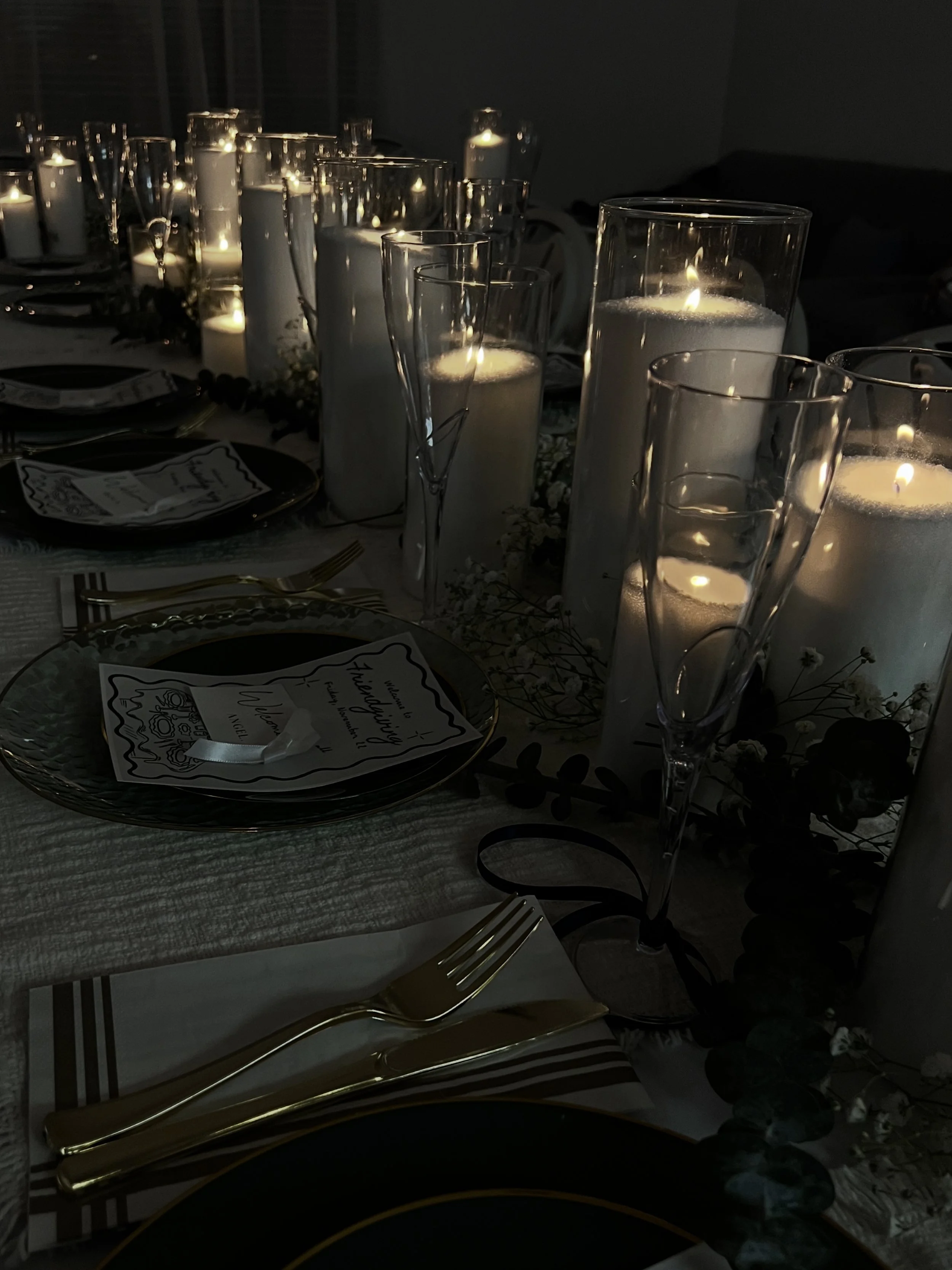 A table set for a celebration with tall glass candle holders containing lit white candles, gold cutlery, black plates, decorative flowers, and place cards.