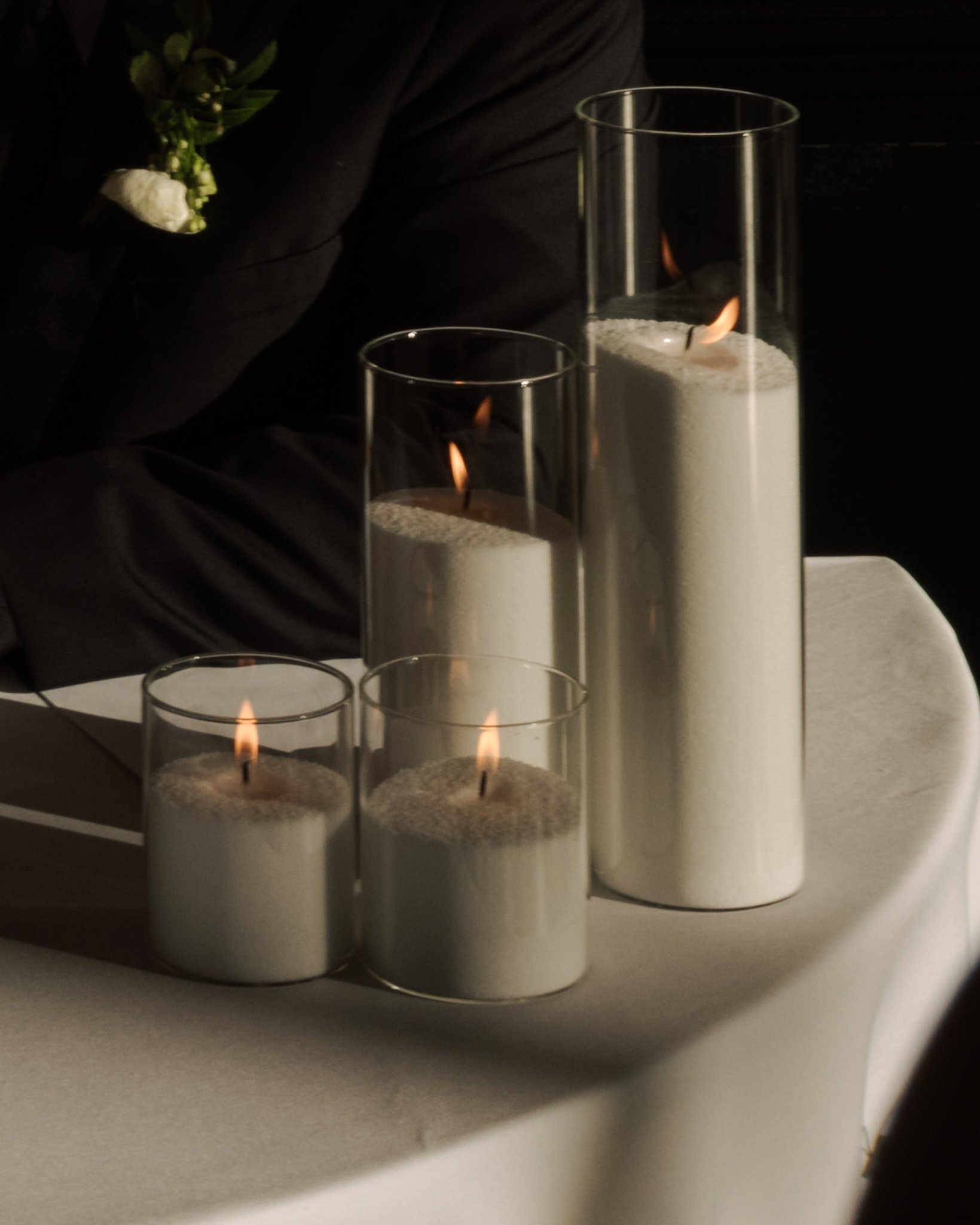 Three lit candles in tall glass holders on a white surface, with a black background and a person's hand and plant in the background.