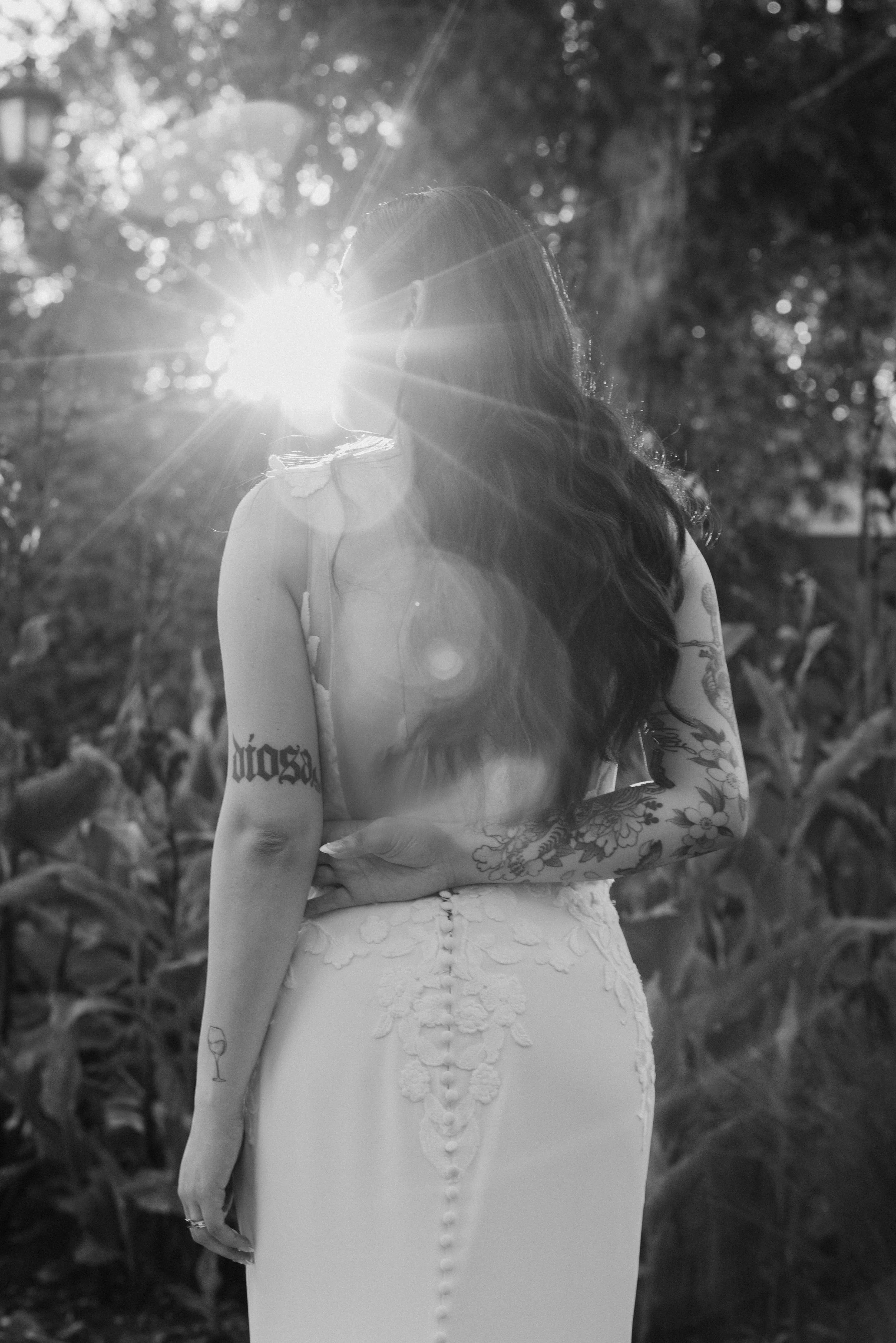 A woman in a white dress standing outdoors with her hand resting on her waist, with the sun shining bright behind her, creating a lens flare effect, and trees in the background.