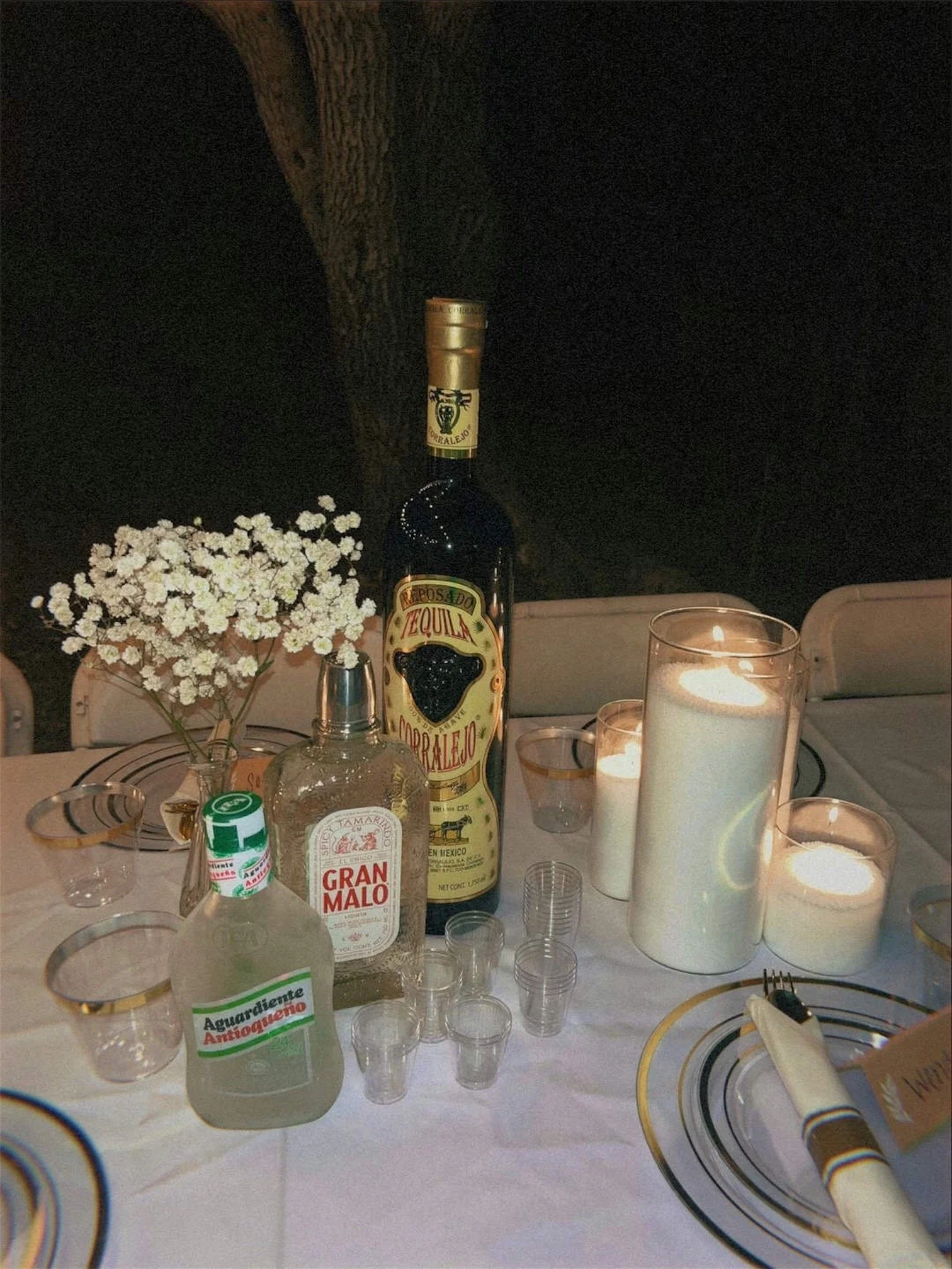 A table set for a celebration with a bottle of tequila, a bouquet of small white flowers, candles, glasses, and tableware.