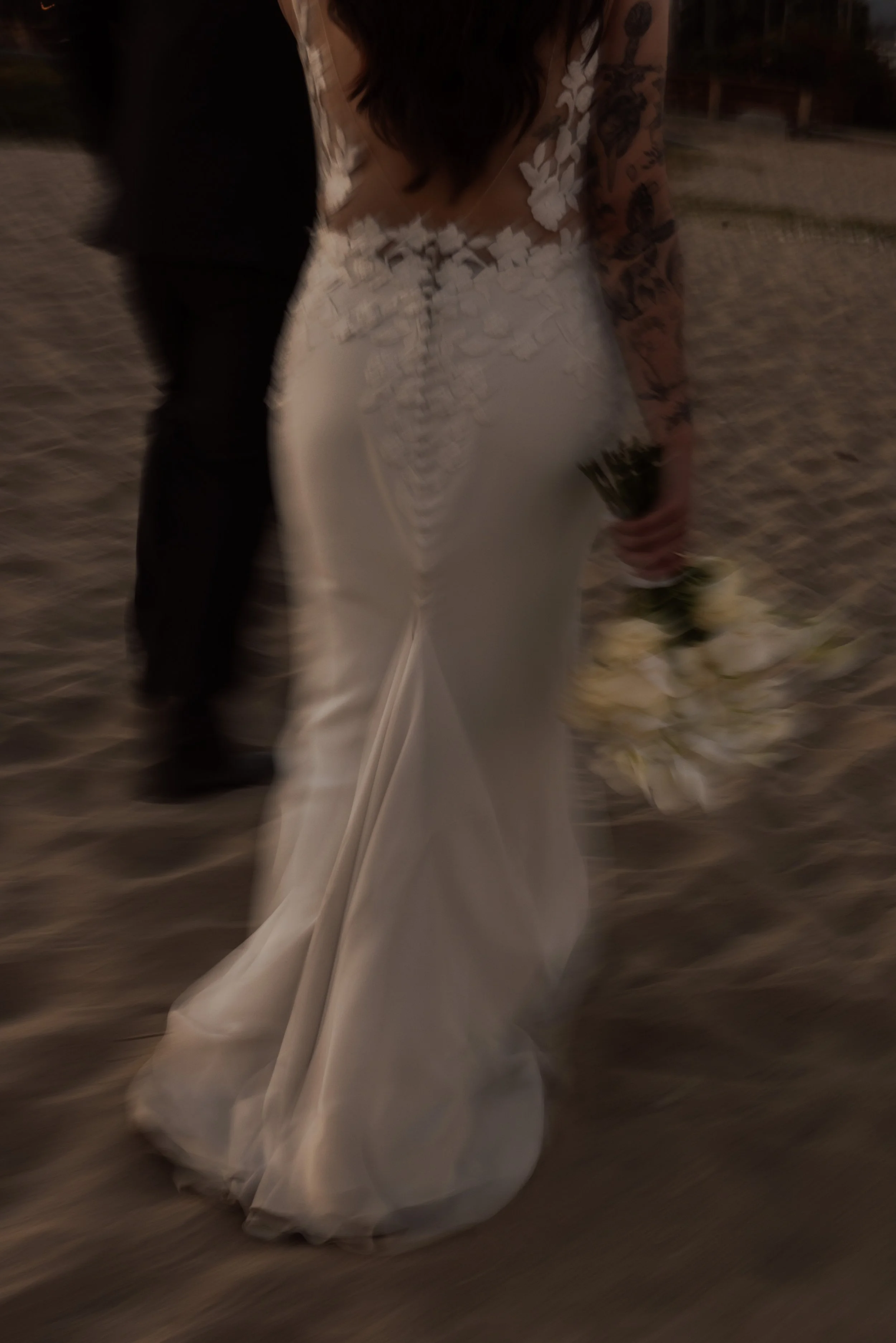 A bride in a white wedding dress with floral embroidery on the top and long train, holding a bouquet of white flowers and standing on a dirt or gravel ground at sunset or evening.