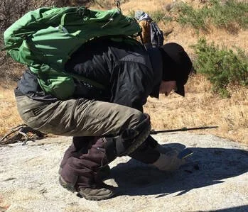 Person crouching outdoors on sandy ground, wearing a green jacket, black hoodie, camo pants, and a black cap, in a sunny area with sparse vegetation.