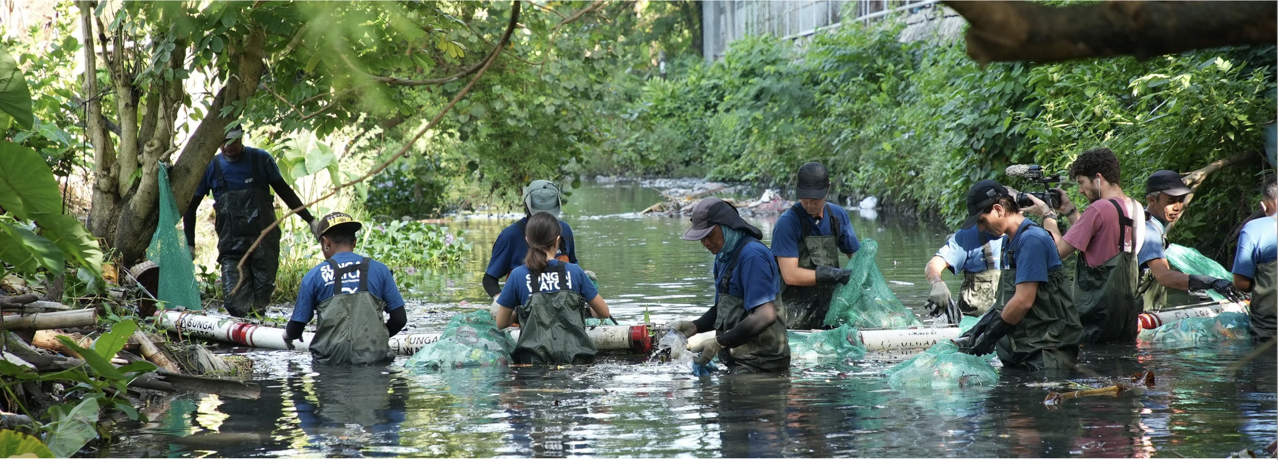 Group of people cleaning a river surrounded by green foliage, wearing gloves and waterproof clothing, some using cameras to document the activity.