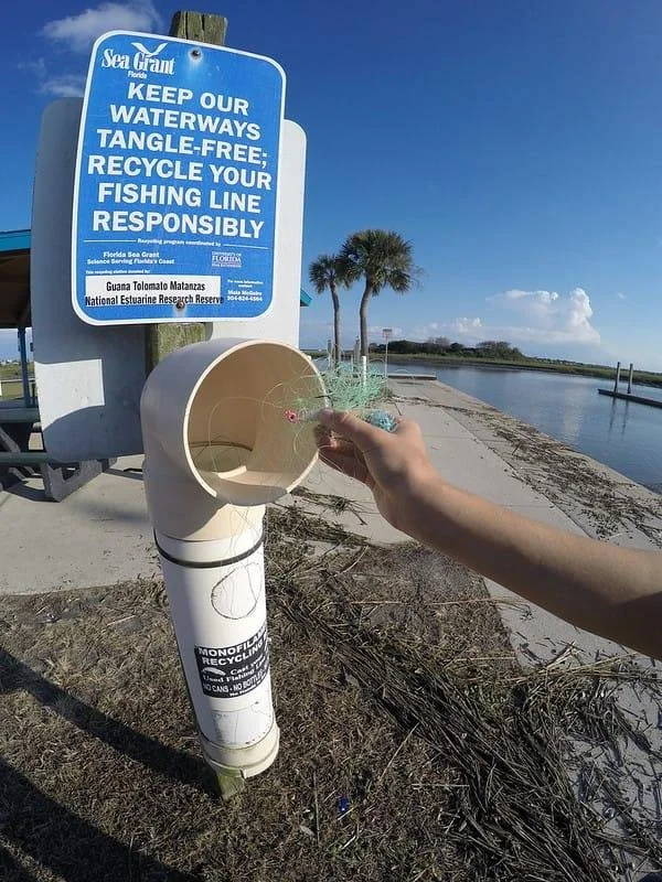 Person's hand placing a fishing line into a designated recycling outlet near a waterway with a blue sign that urges keeping waterways tangle-free and recycling fishing line responsibly, with palm trees and a body of water in the background.