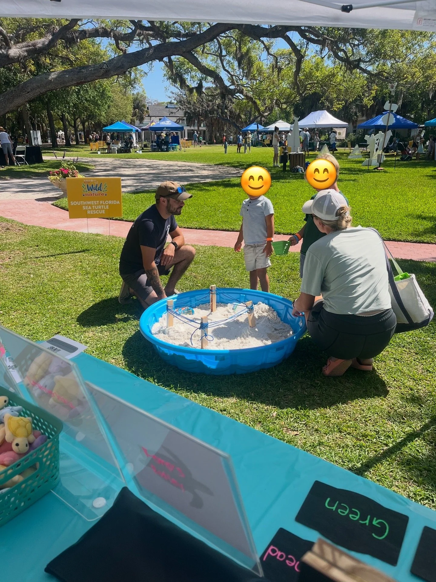 A park scene with multiple tents at an outdoor event. In the foreground, there is a small sand table with toy figures, and a person kneeling next to it. Two children with emoji faces are standing near the sand table, engaging with the toys, while an adult crouches nearby. A sign indicates the event is organized by Southwest Florida Sea Turtle Rescue. The background shows more tents, people, and large trees providing shade.