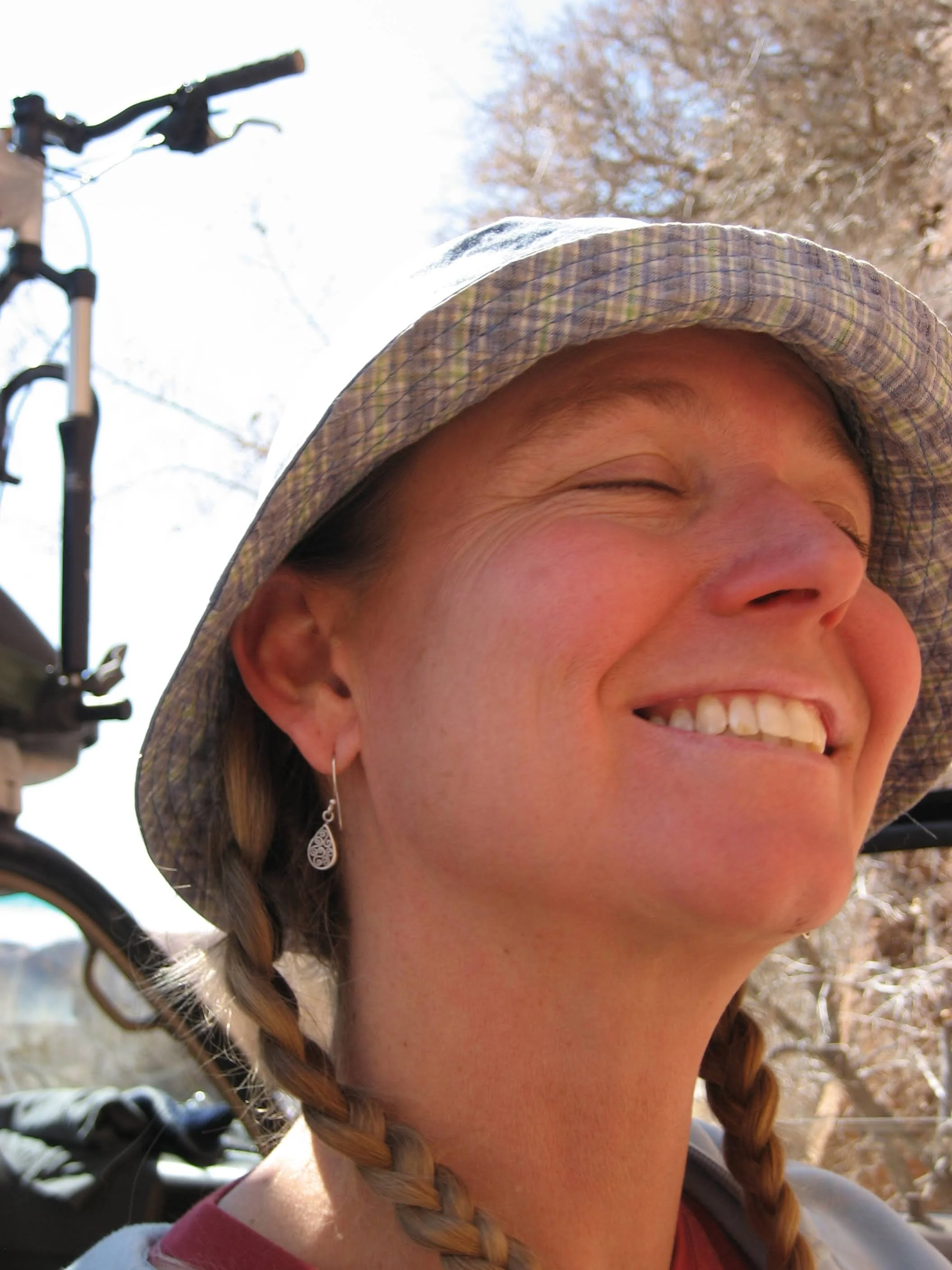 Close-up of a smiling woman with braided hair wearing a bucket hat outdoors, with a bicycle and trees in the background.