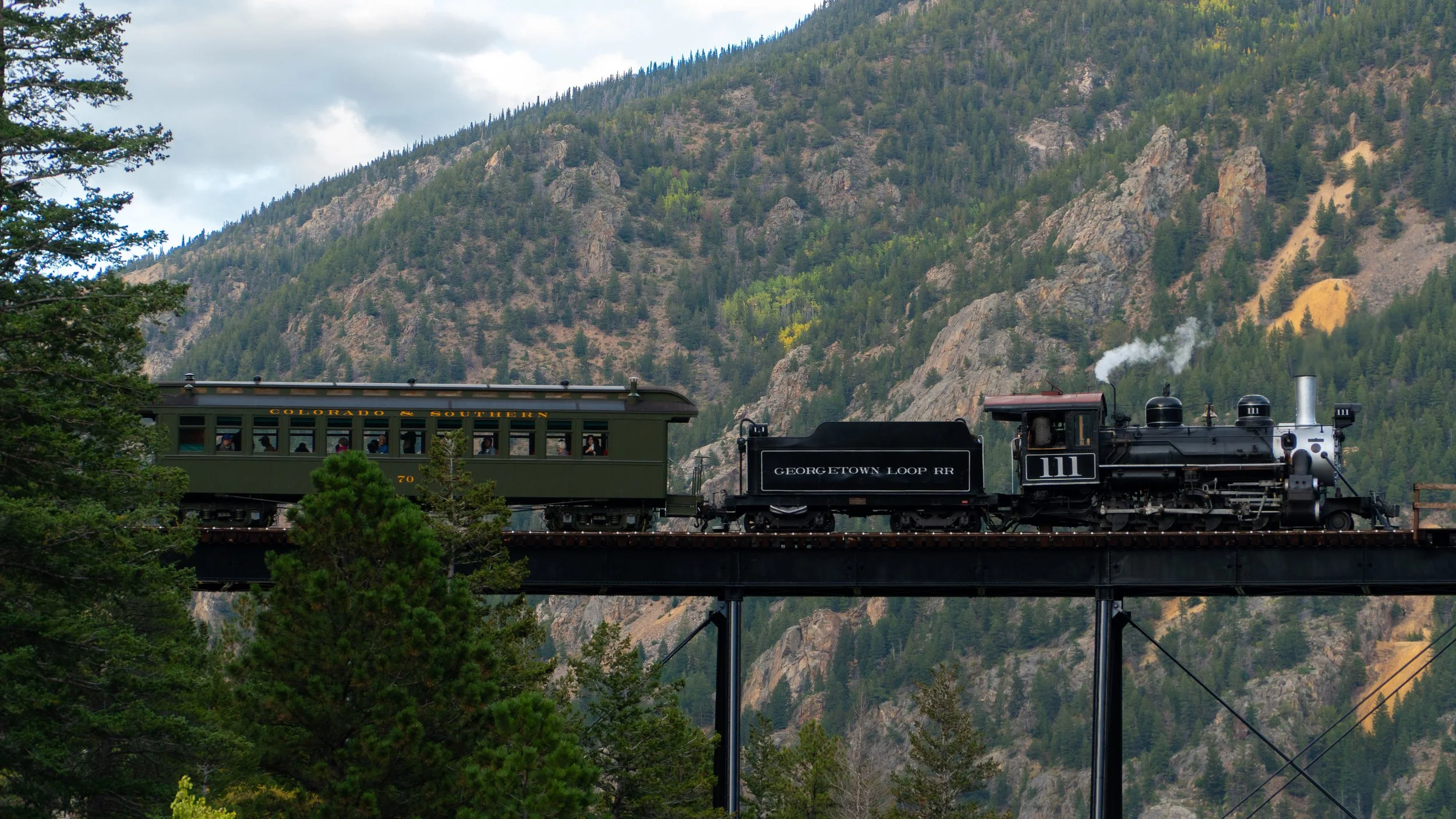A vintage steam train with a black locomotive and a green passenger car traveling across a bridge through a mountainous forested area, with tall pine trees and rocky hillside in the background.