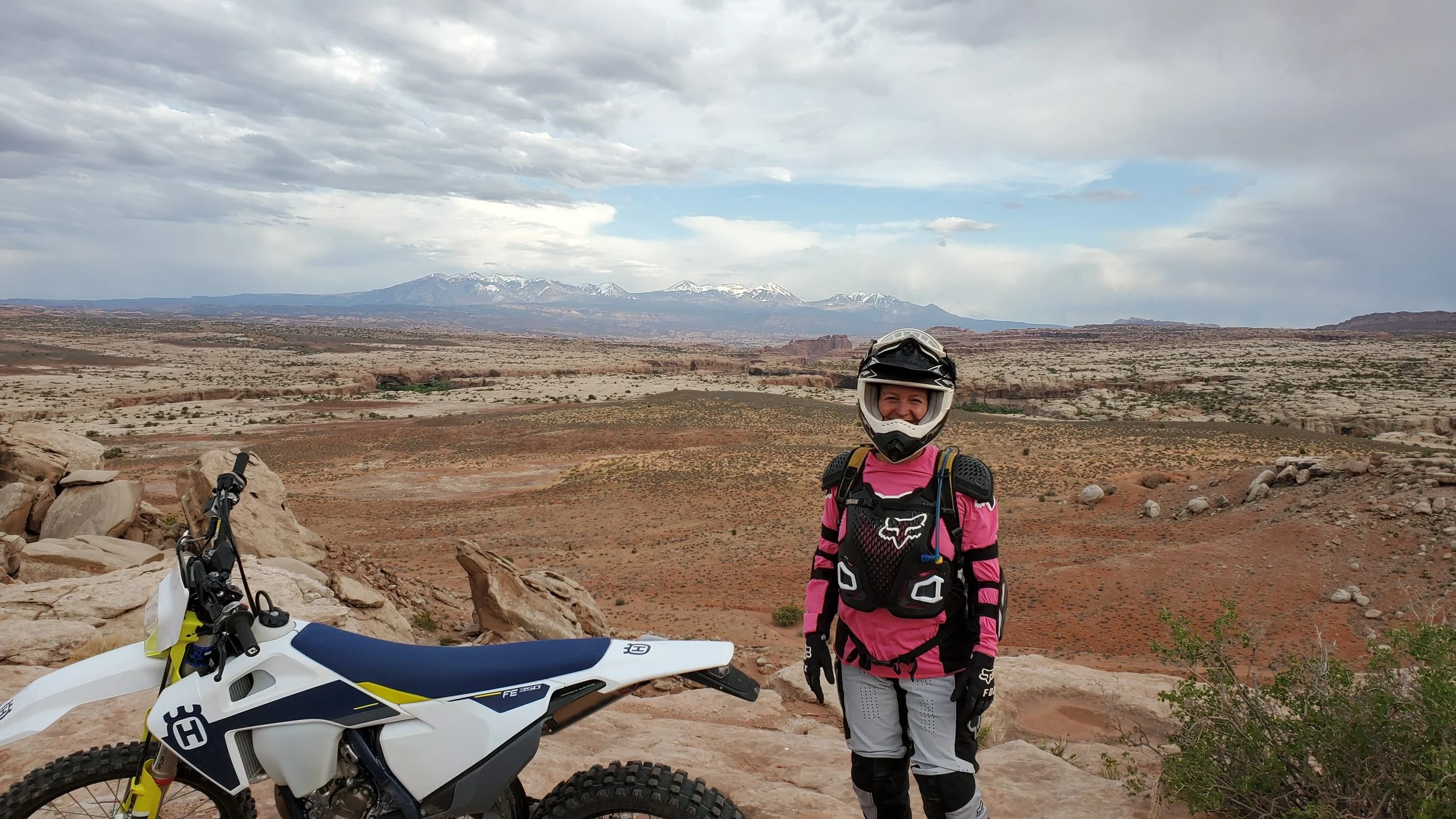 A person wearing a pink and black motocross outfit and a helmet, standing next to a white dirt bike on a rocky hilltop in a desert landscape with mountains in the background.