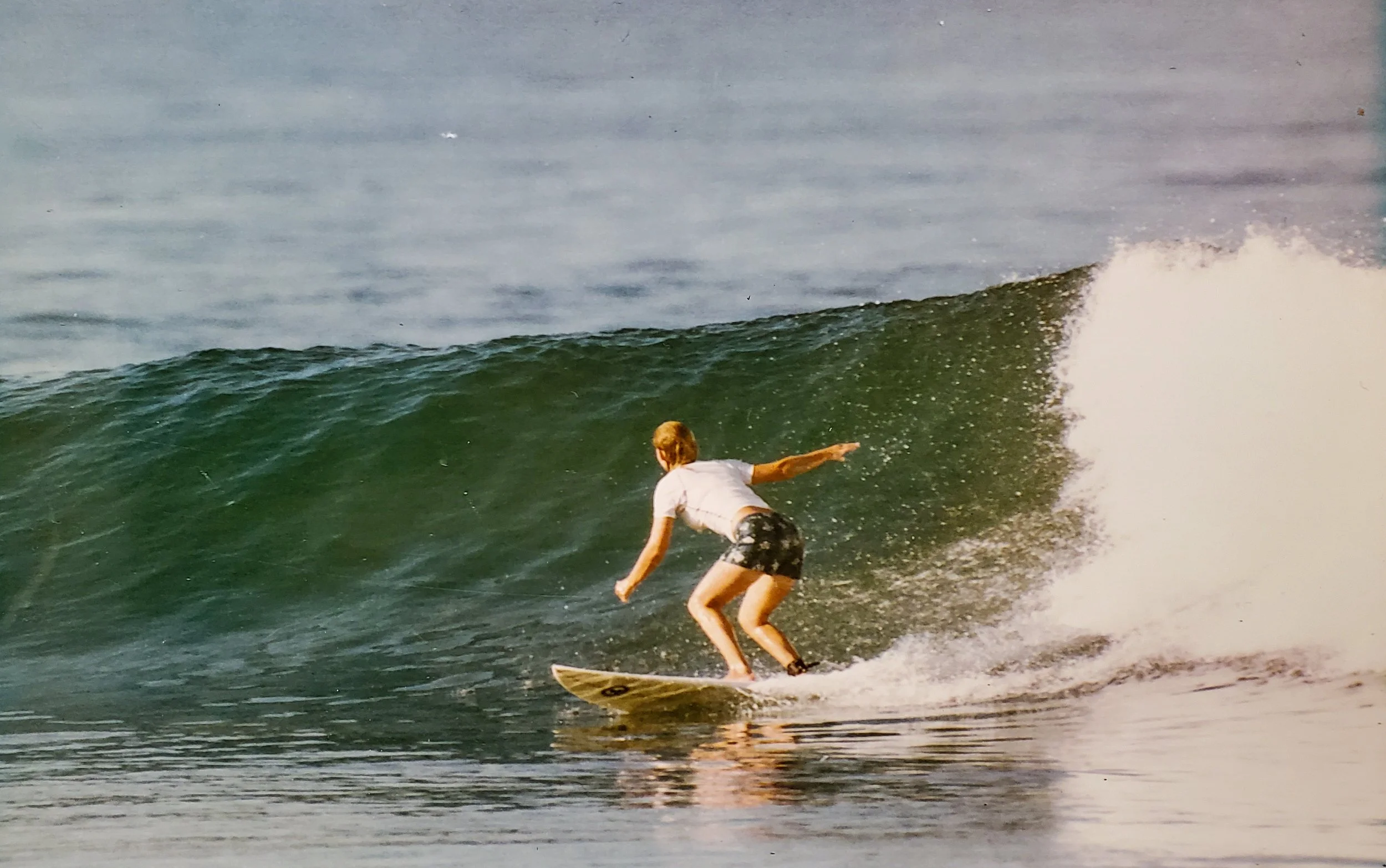 A person with blond hair wearing a white t-shirt and dark patterned shorts riding a surfboard on a green wave in the ocean.