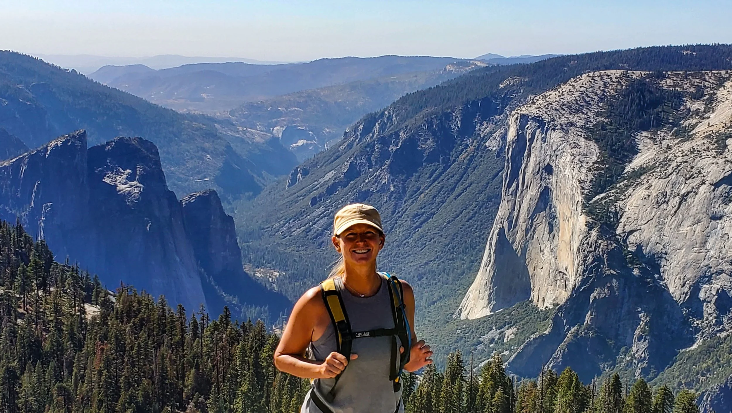A woman hiking in a mountainous area with steep cliffs, forests, and distant mountain ranges.