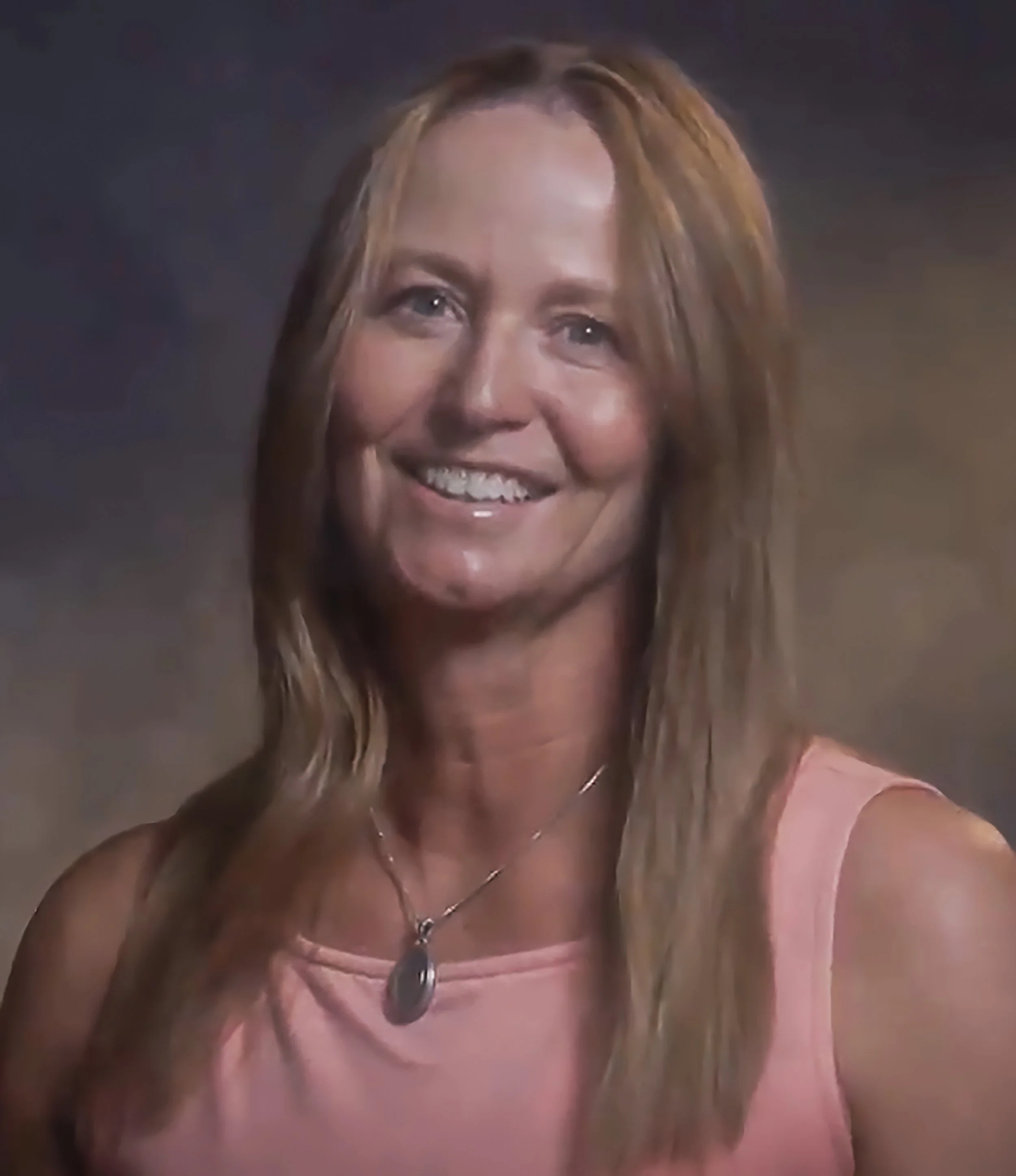 A woman smiling. She is wearing a pink top and a pendant necklace.