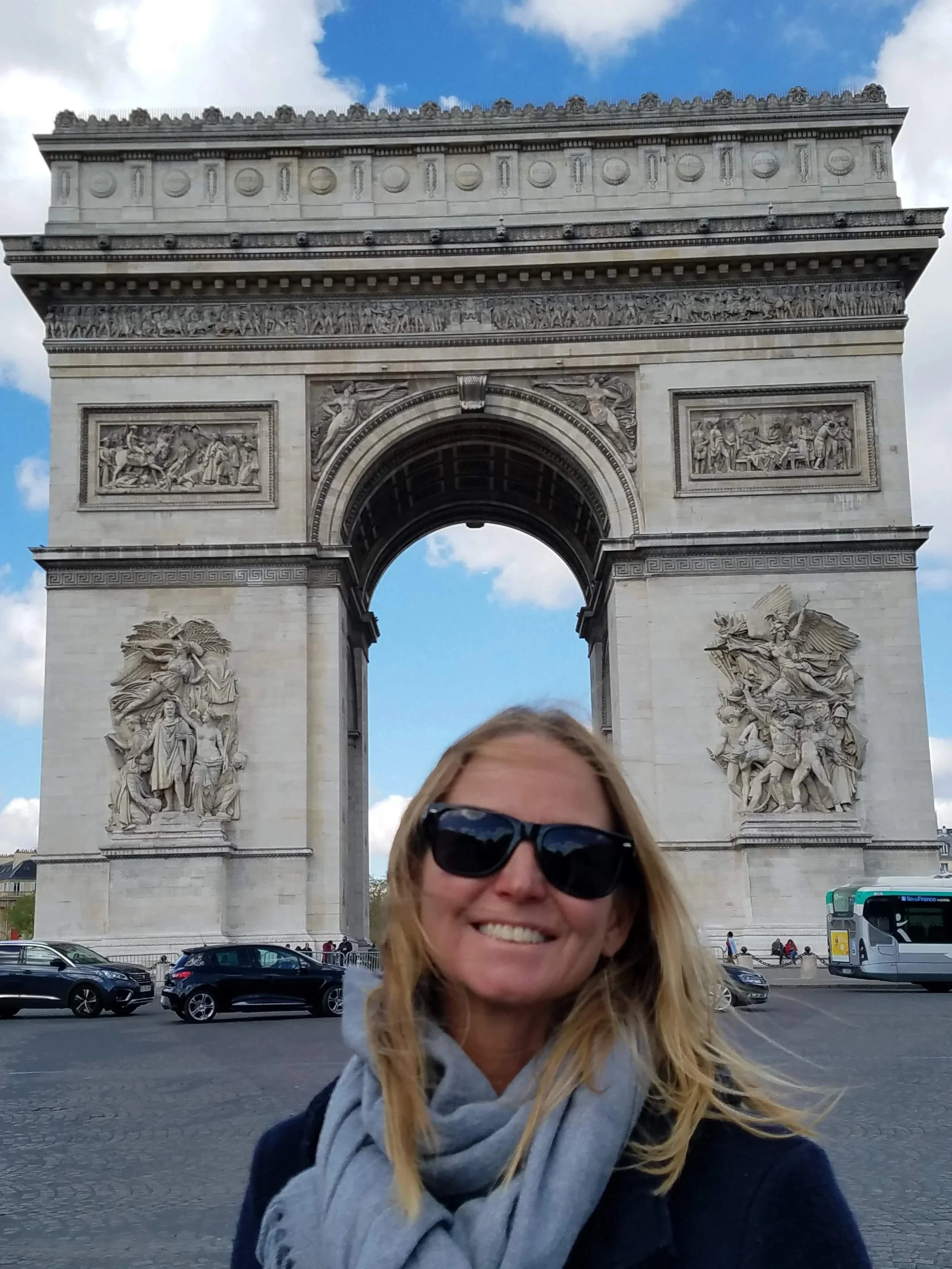 Person with blonde hair and sunglasses smiling in front of the Arc de Triomphe in Paris, France.