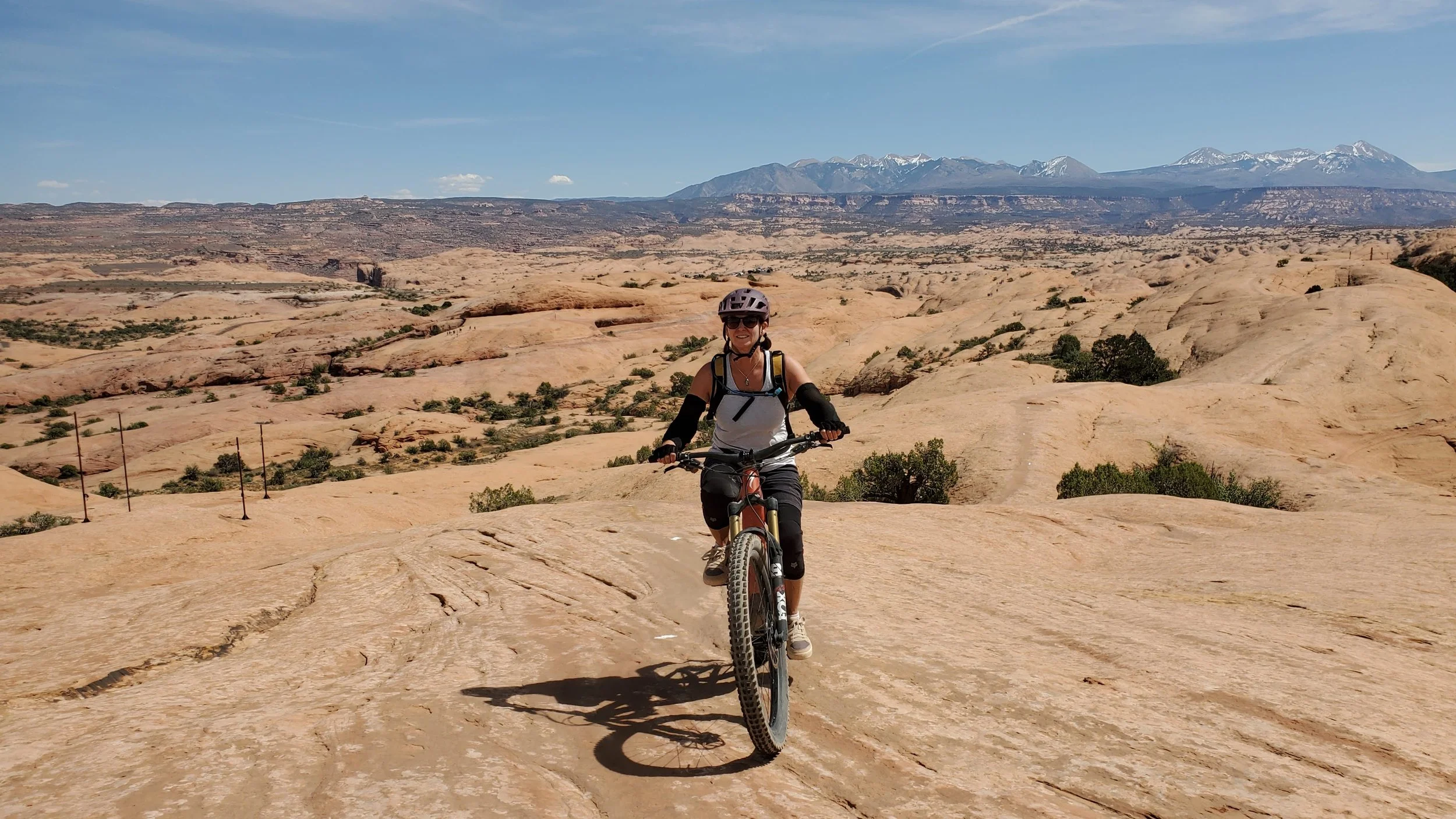 A woman in biking gear, including a helmet, sunglasses, and gloves, rides a mountain bike on a rugged, arid landscape with large rock formations. In the background, there are distant mountains with snow-capped peaks under a blue sky.