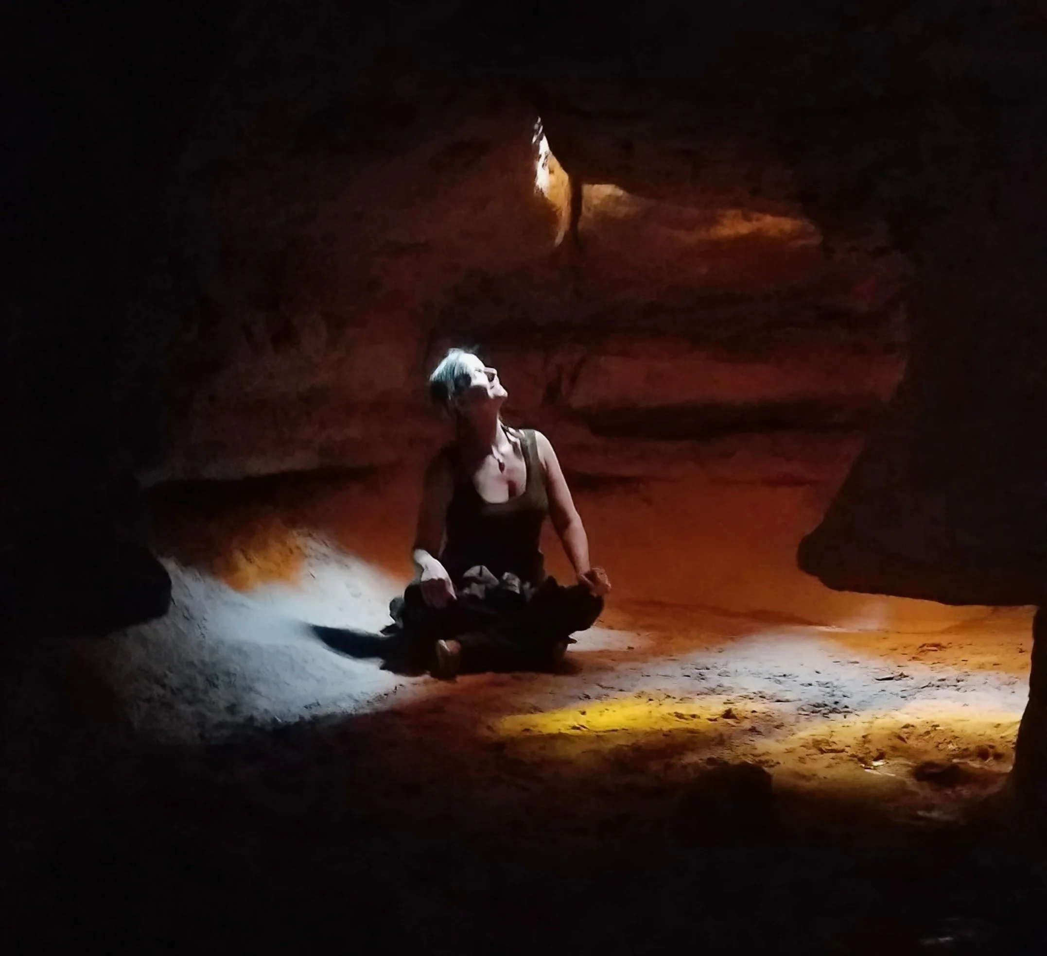 A woman sitting inside a dark cave or tunnel, illuminated by a dim light coming from the entrance, with a rugged, rocky interior.