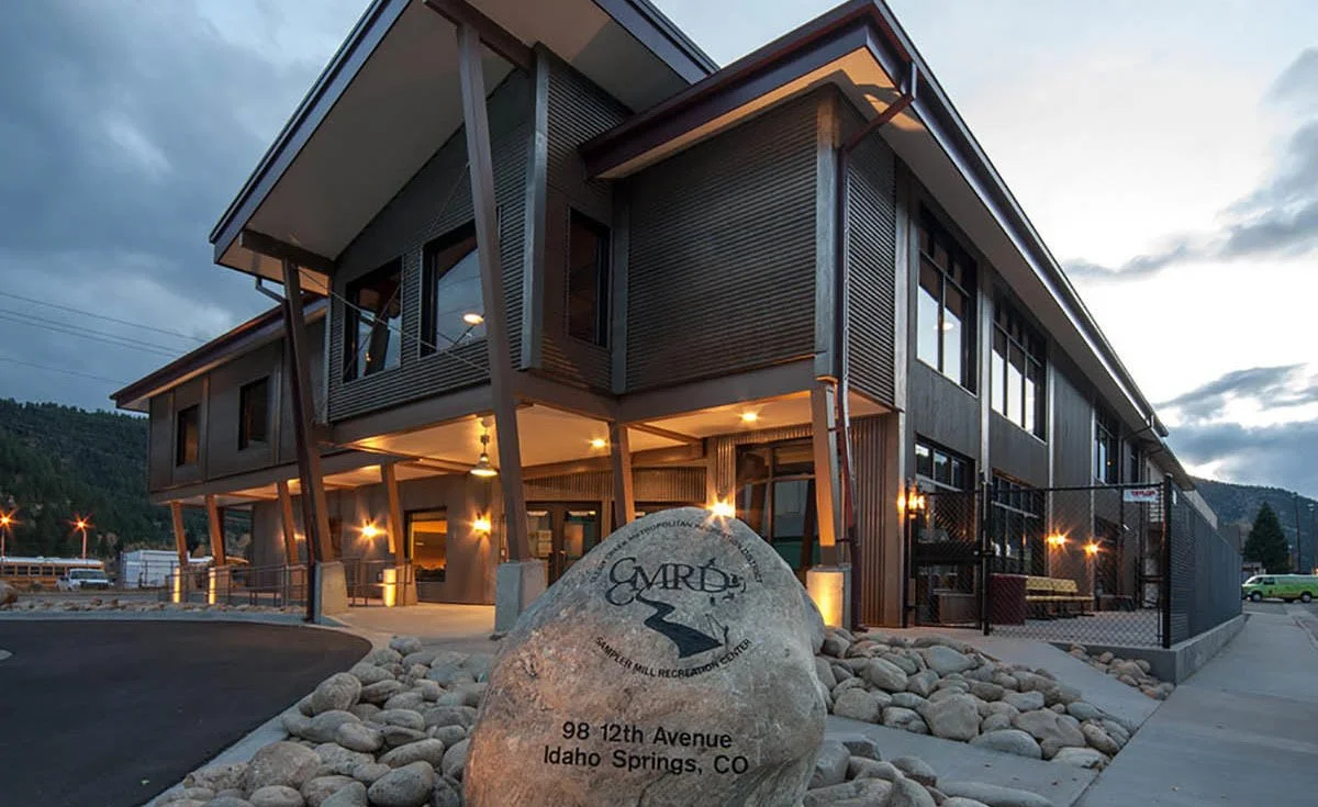 Exterior of a modern building with large windows, metal siding, and angled support beams, surrounded by rocks and a parking lot, with a sign indicating the location in Idaho Springs, Colorado.