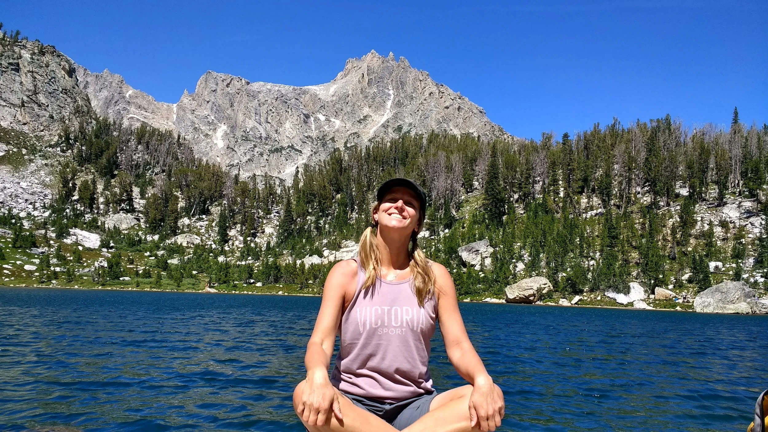 A smiling woman sitting cross-legged on a rock near a large lake, with a mountainous landscape and green trees in the background under a clear blue sky.