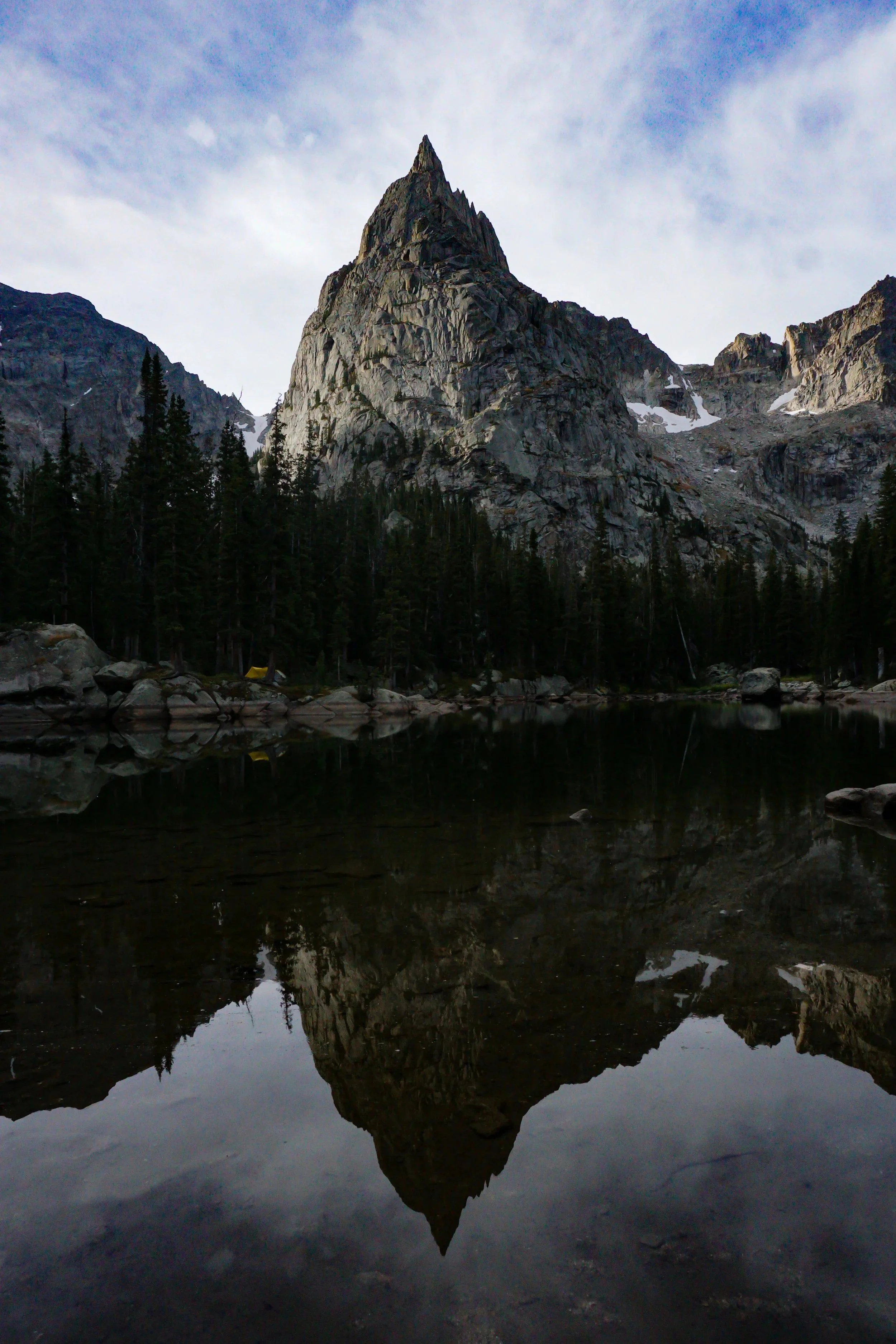 A tall, pointed mountain with rocky slopes is reflected in a calm mountain lake, surrounded by dark green trees under a partly cloudy sky.