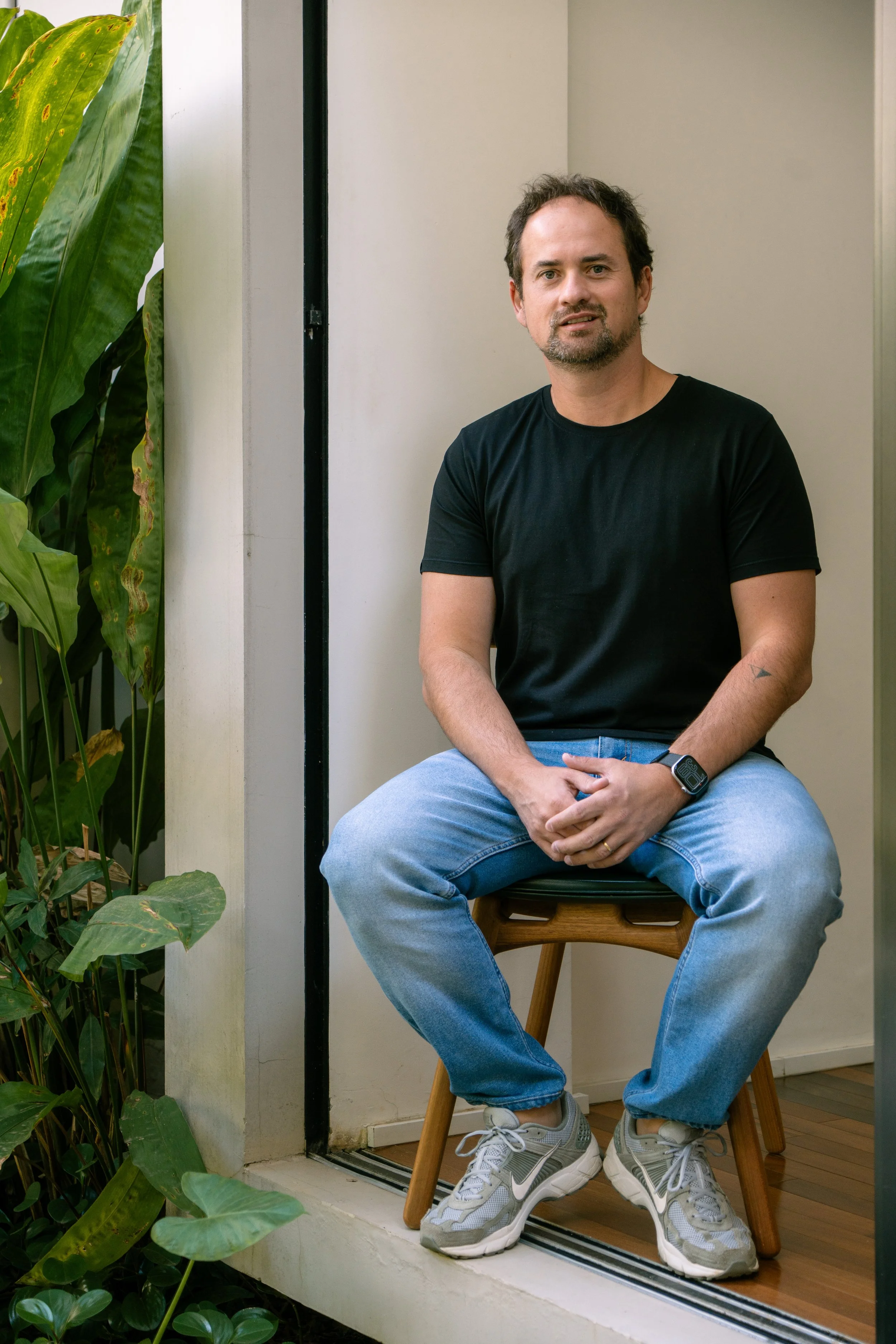 Man sitting on a wooden chair in a room, with green plants on the left.